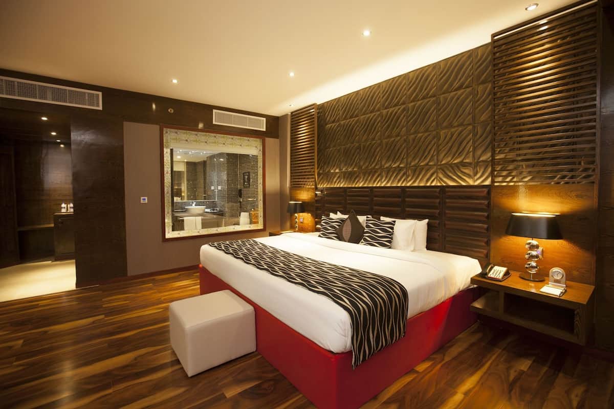 A modern hotel room with a large bed, a bedside table with a lamp, a mirror, and a wooden floor.
