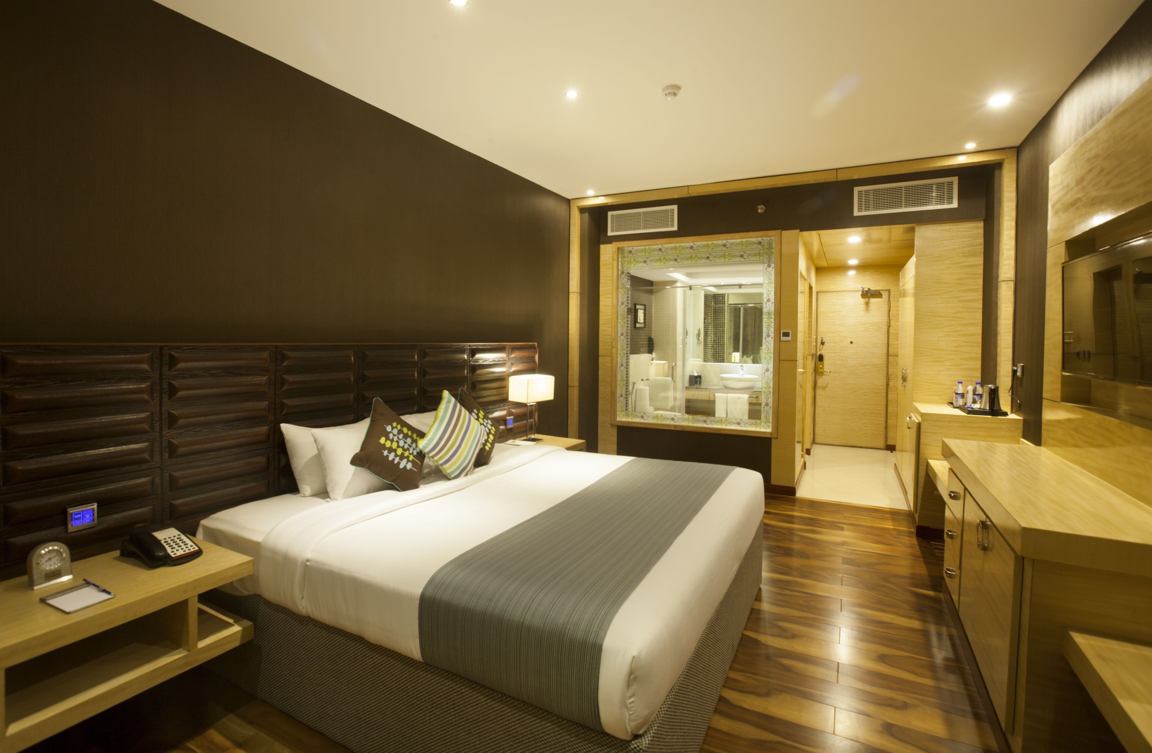 Una habitación de hotel moderna con una cama grande, una mesita de noche y un escritorio pequeño. Hay un baño con espejo y lavamanos.