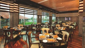 A spacious restaurant with multiple dining tables and chairs, large windows offering a view of the outdoors, and a modern interior design. - Ramada by Wyndham Alleppey (Ambalapuzha)