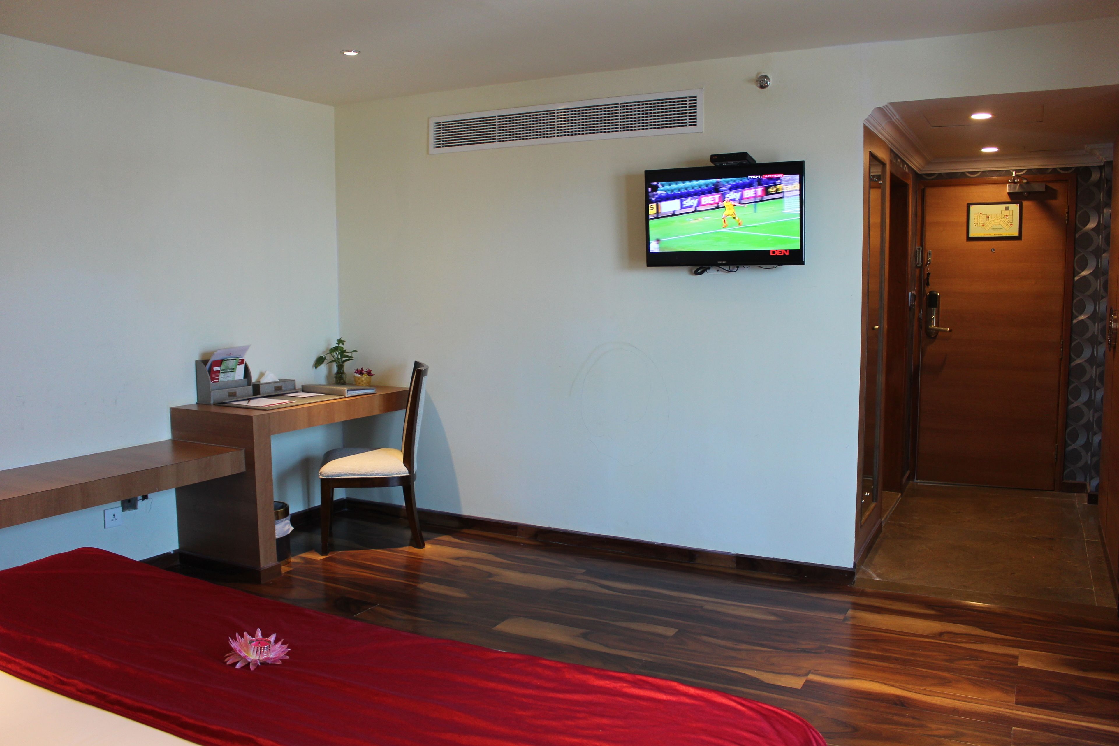 A hotel room with a wooden desk, a chair, a TV mounted on the wall, and a red bedspread.