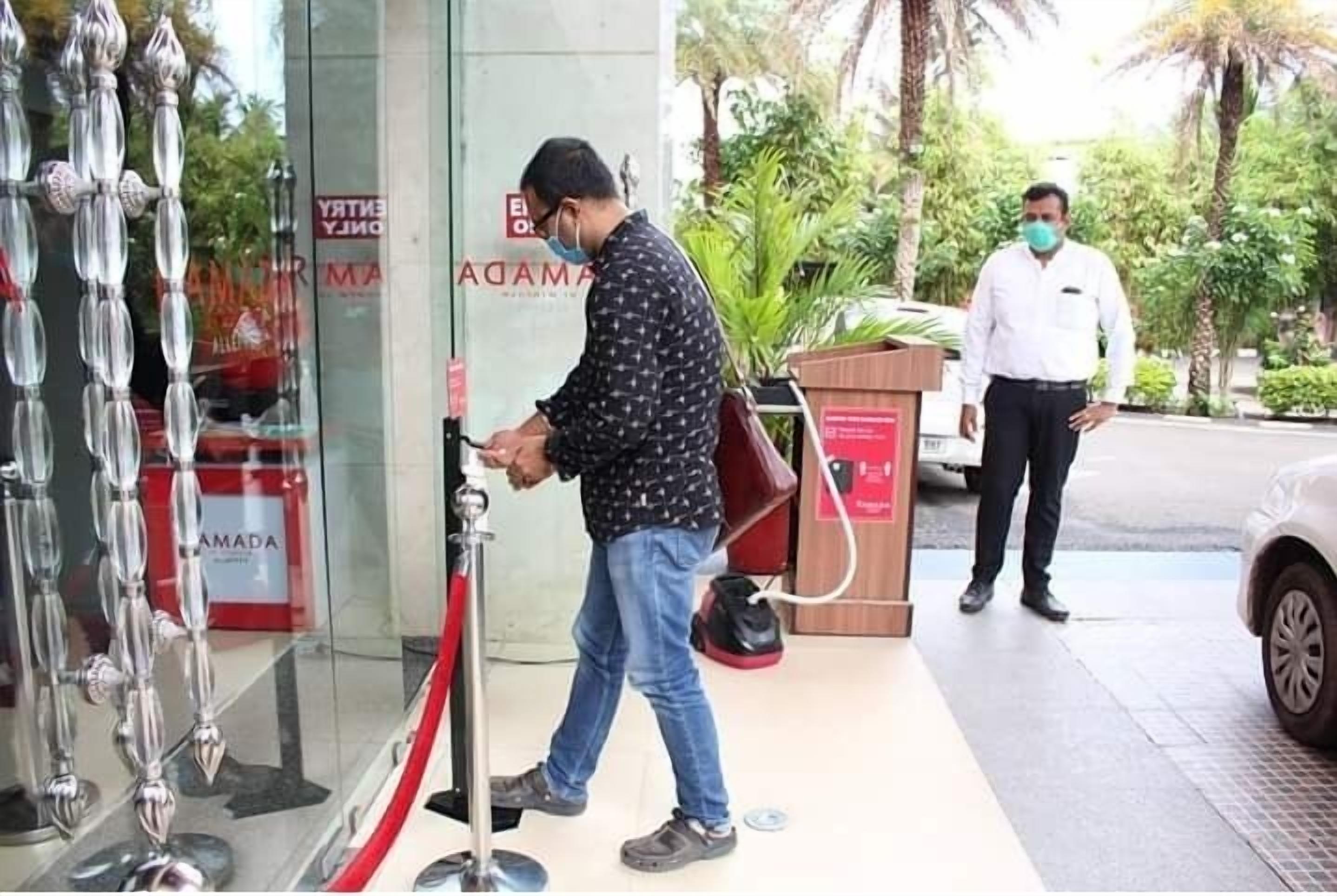 A man walking through a hotel entrance with a red rope barrier, a sign reading "ADAMA," and a man in a white shirt standing nearby.