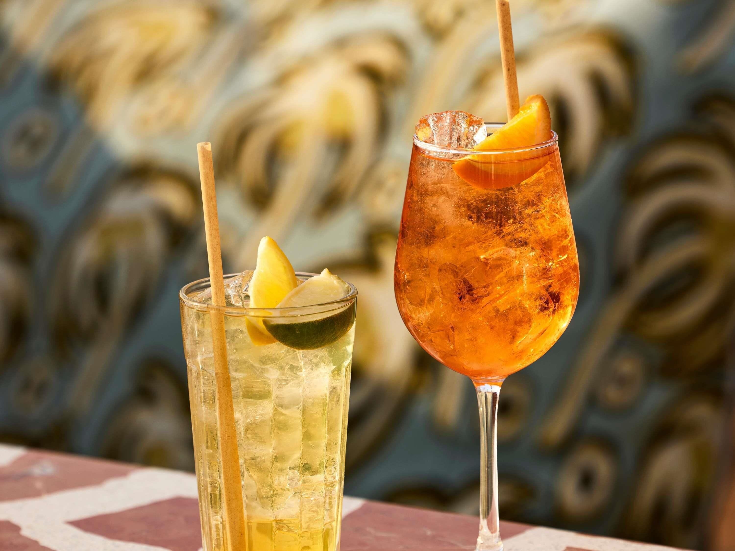 Two drinks with straws and fruit slices, one in a short glass and one in a tall glass, on a table with a patterned background.