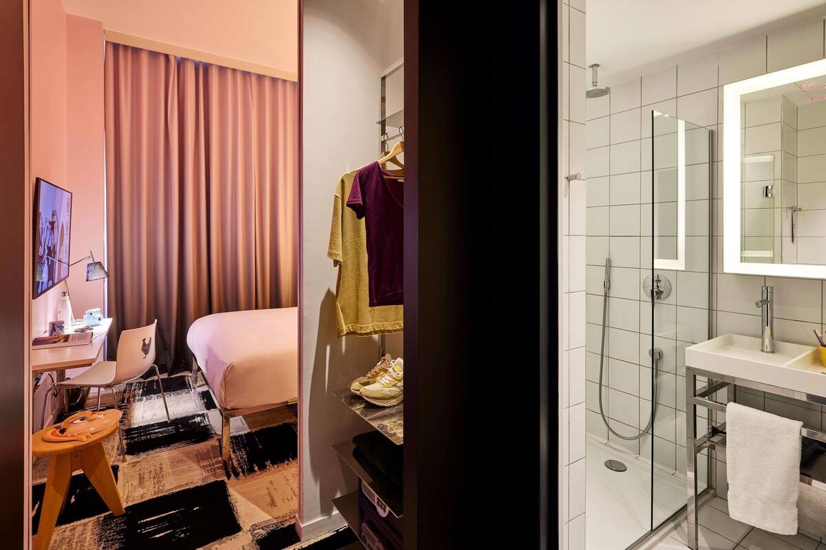 Hotelkamer met een bed, een bureau met stoel, een televisie, een klein krukje en een badkamer met douche, wastafel en spiegel.