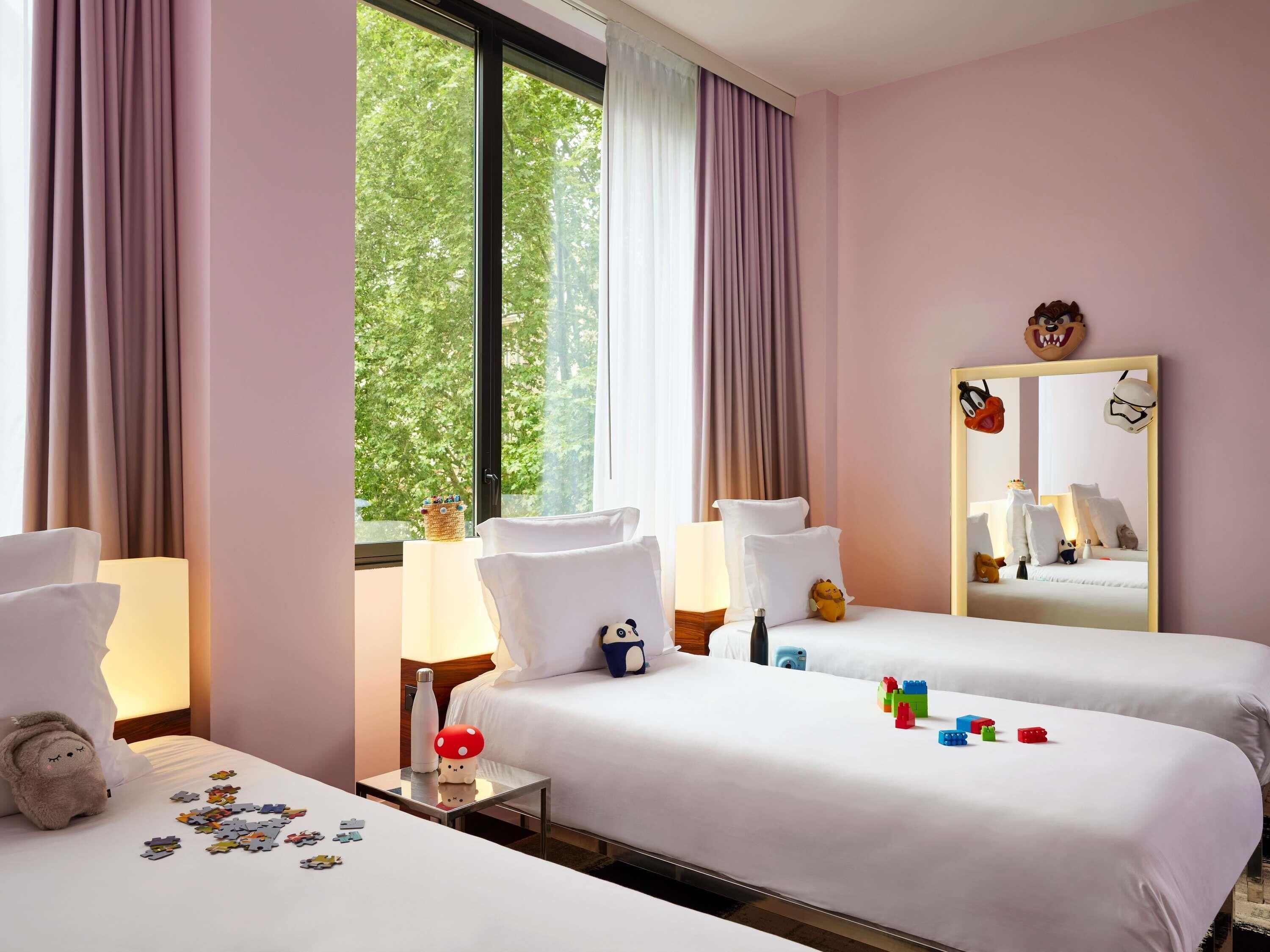 A hotel room with two beds, a large window with curtains, a bedside table with a lamp, a mirror, and a stuffed toy on one bed.