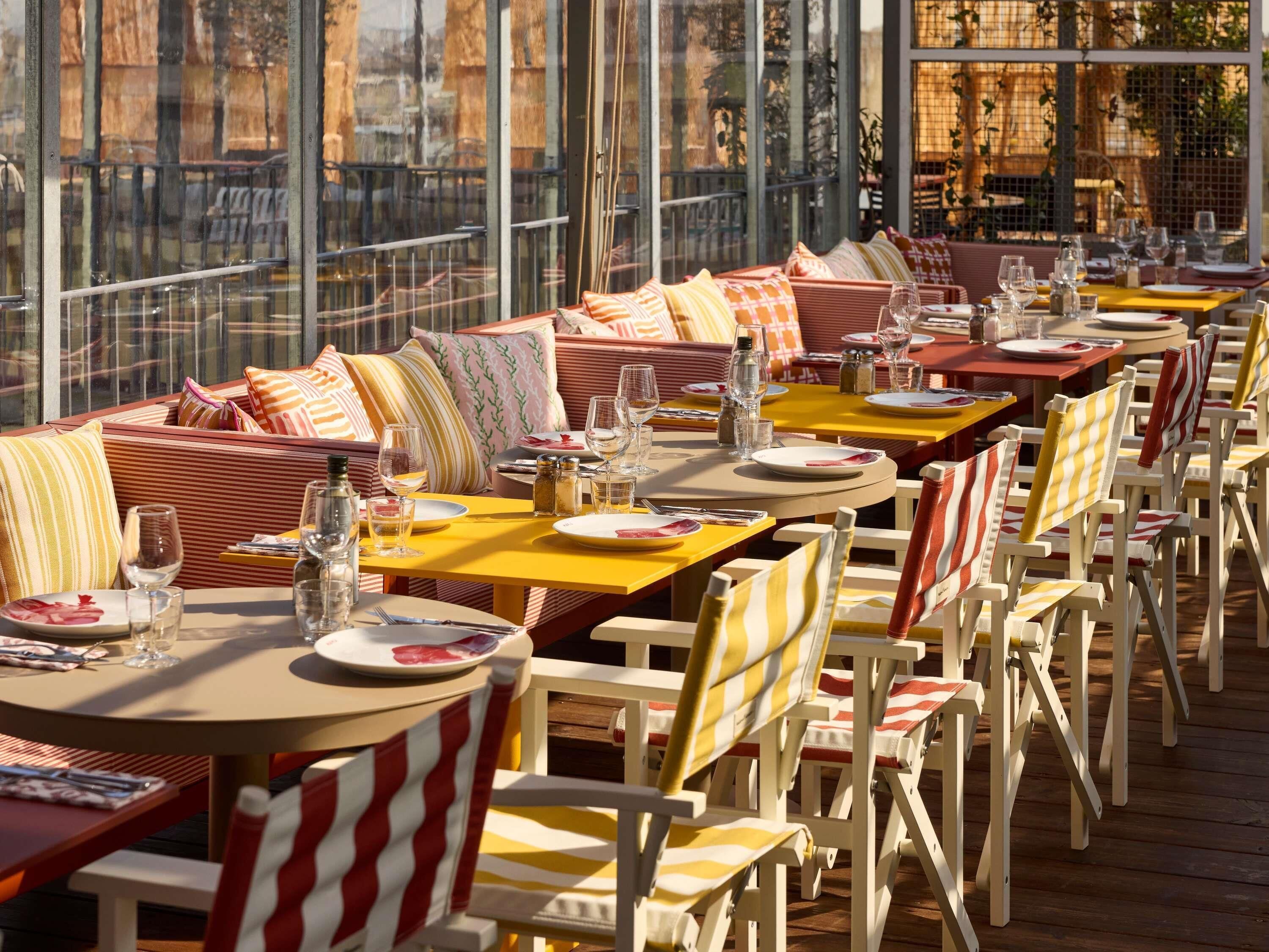 An outdoor dining area with tables and chairs, large windows, and a view of the city.