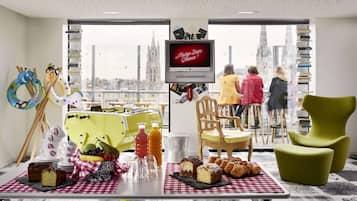 Quarto com uma mesa posta com doces e bebidas, uma televisão exibindo "Mama Says Cheese!", e vista para um horizonte urbano através de grandes janelas.