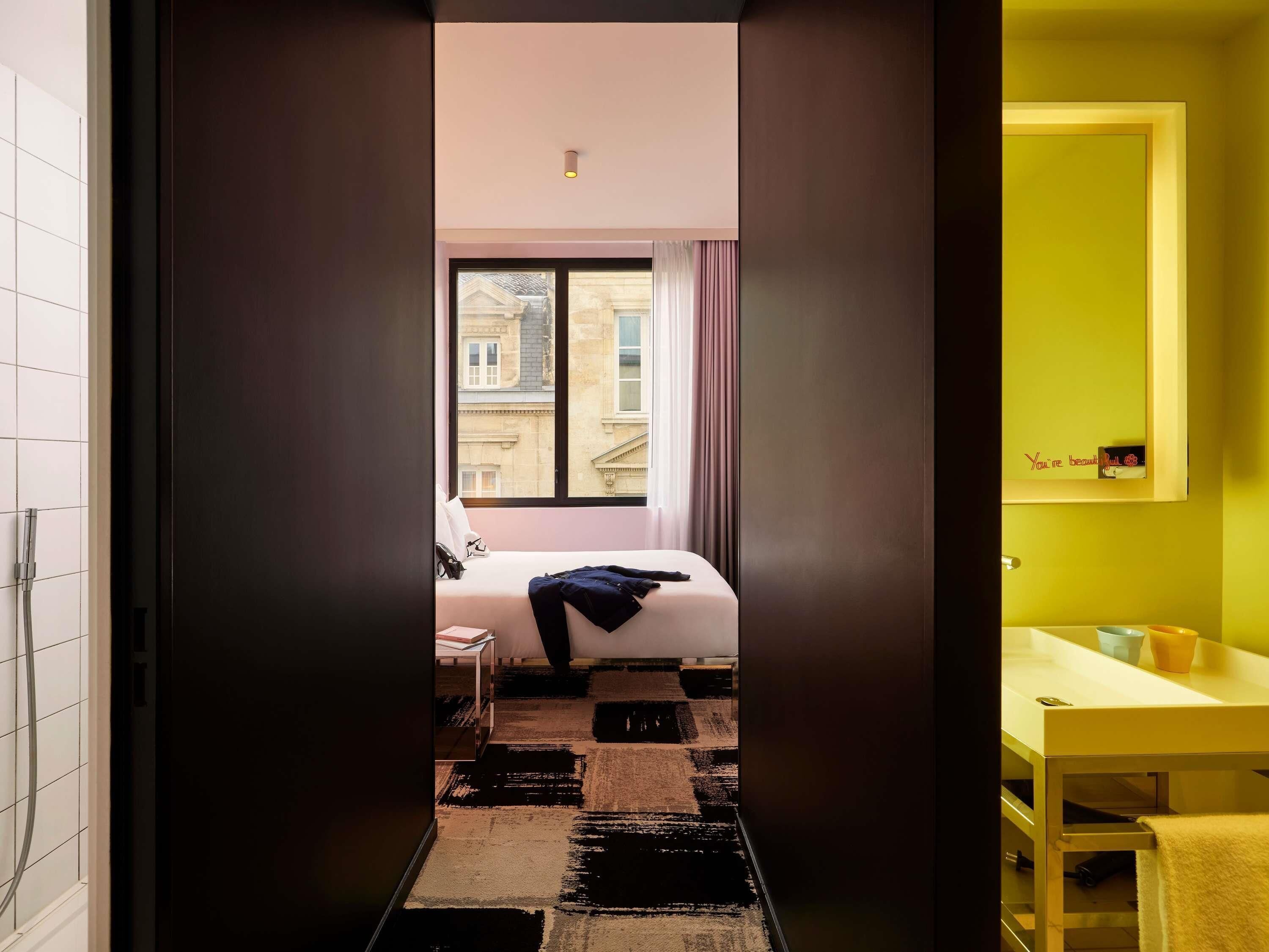 Una habitación de hotel con una cama, un ventanal con vistas a edificios, un baño con pared amarilla y una alfombra de estampado blanco y negro.