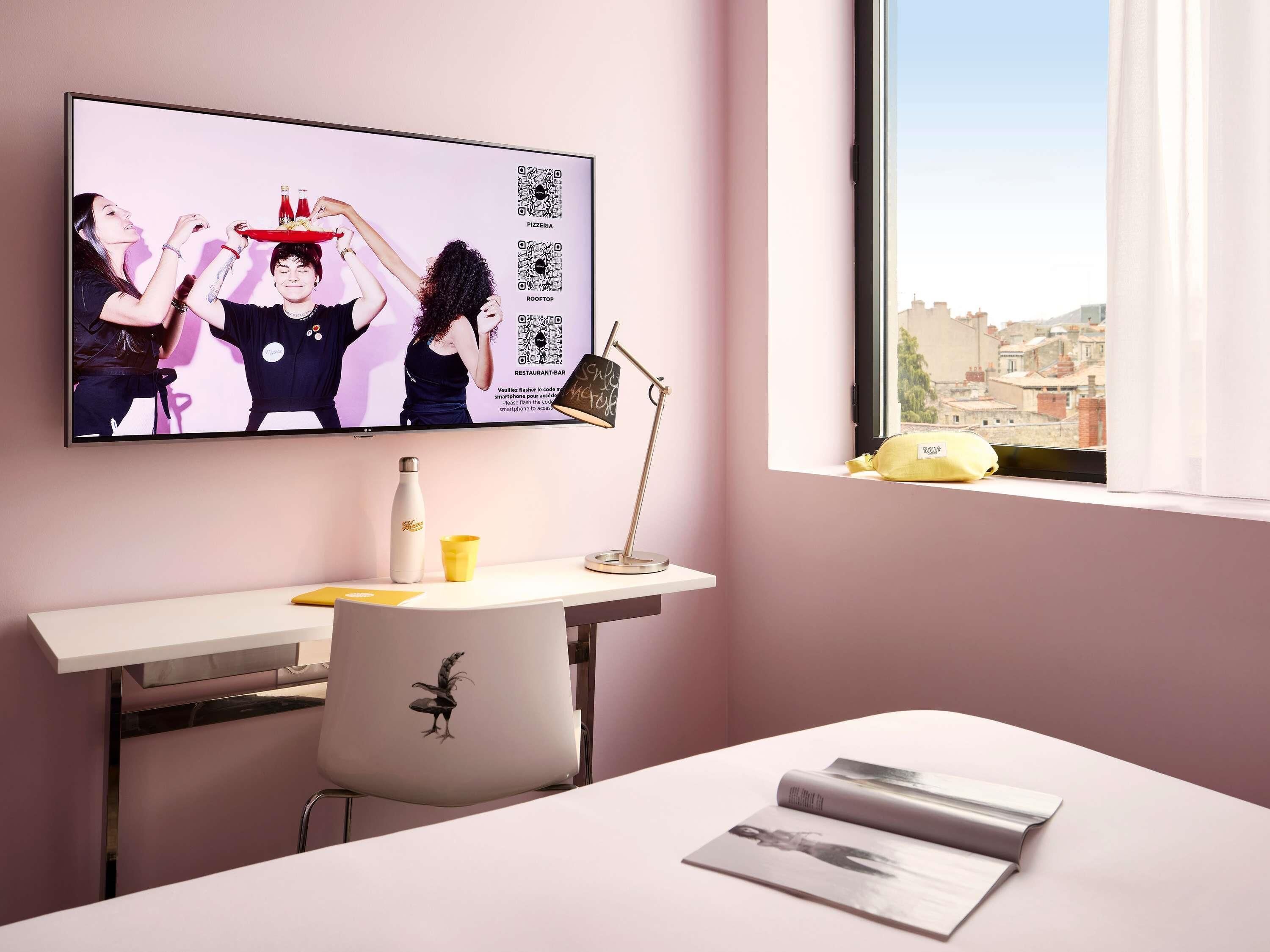 A modern room with a flat-screen TV, a desk with a chair, a window with a view of buildings, and a small table with a water bottle and a cup.