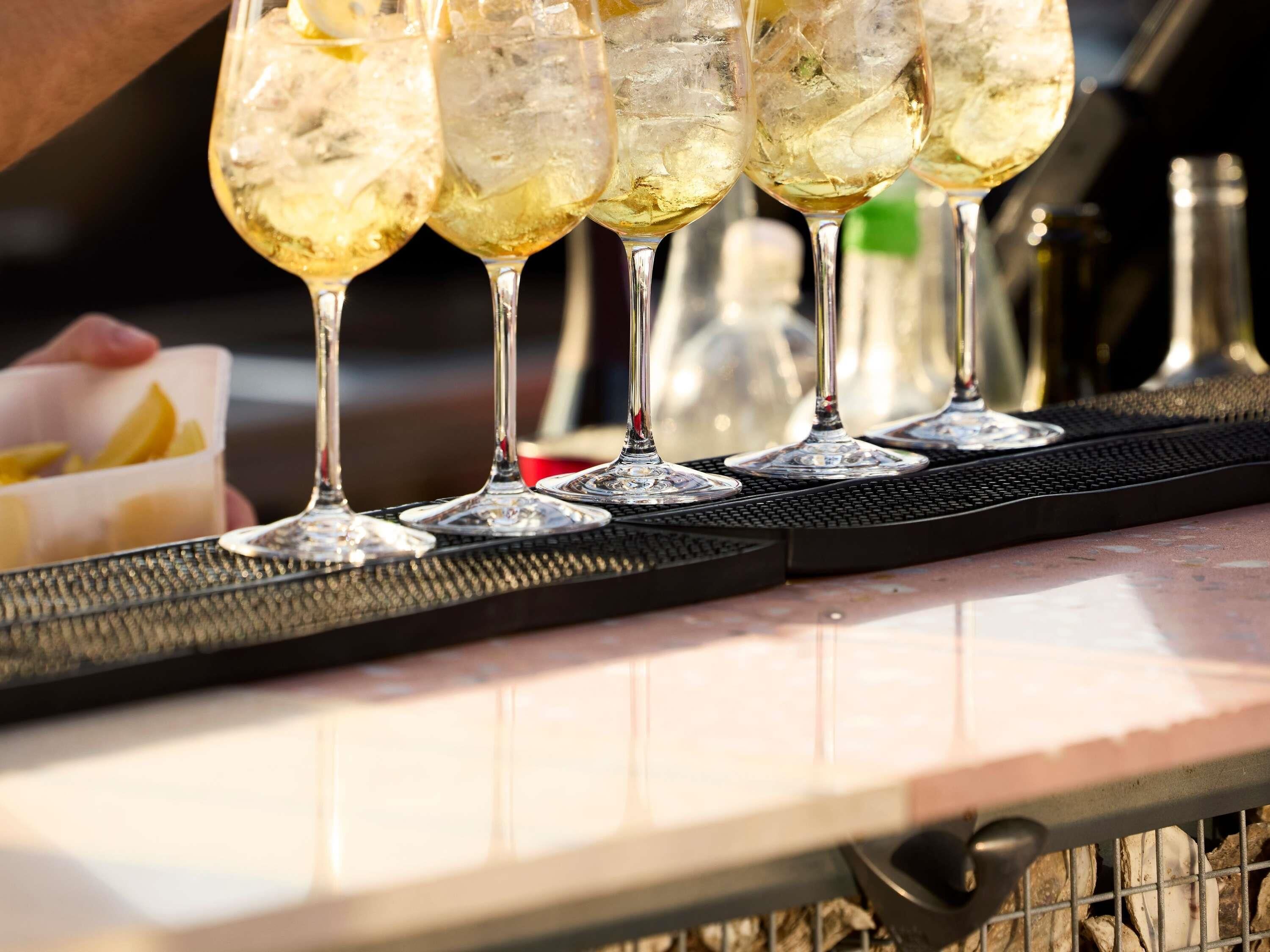 A bar setting with glasses of a clear beverage, a black bar mat, and a container with lemon slices.