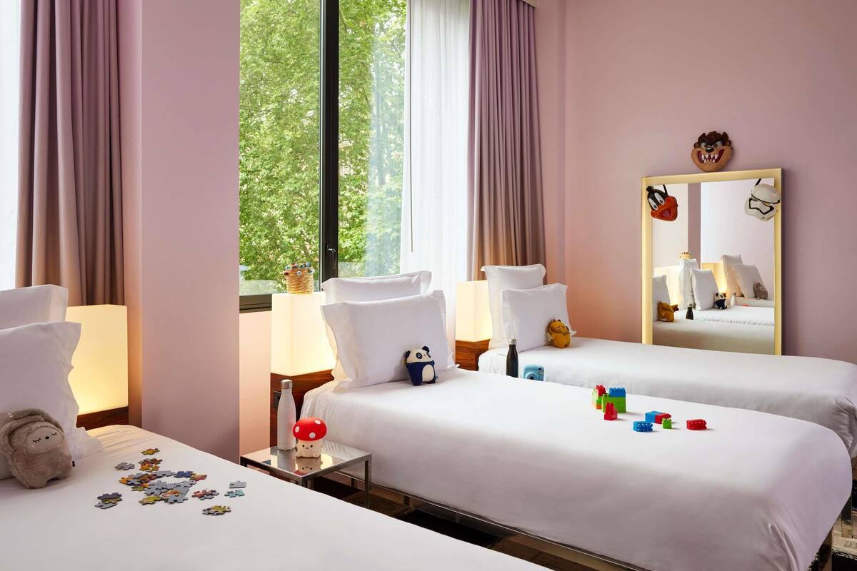 Hotelkamer met twee bedden, een groot raam met gordijnen, een spiegel en een klein tafeltje met een lamp en een pluchen speelgoed.