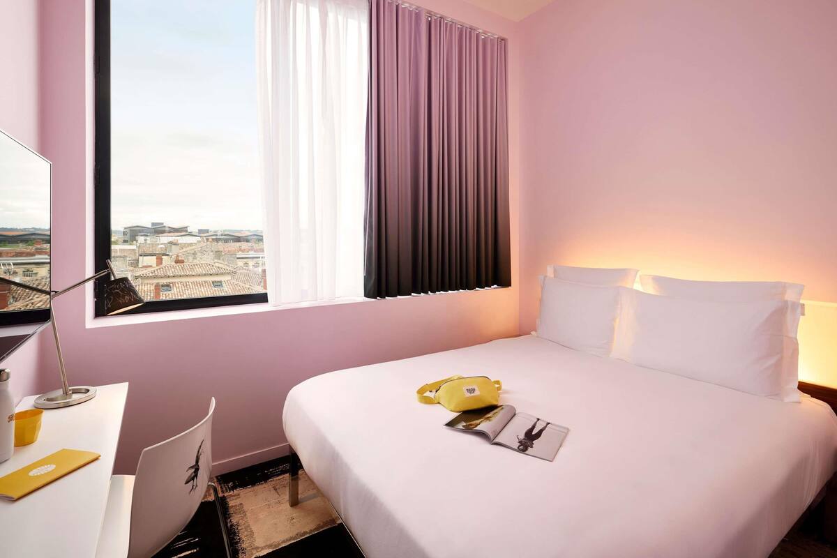 Hotelkamer met een bed, een bureau, een stoel, een raam met gordijnen en uitzicht op de stad.