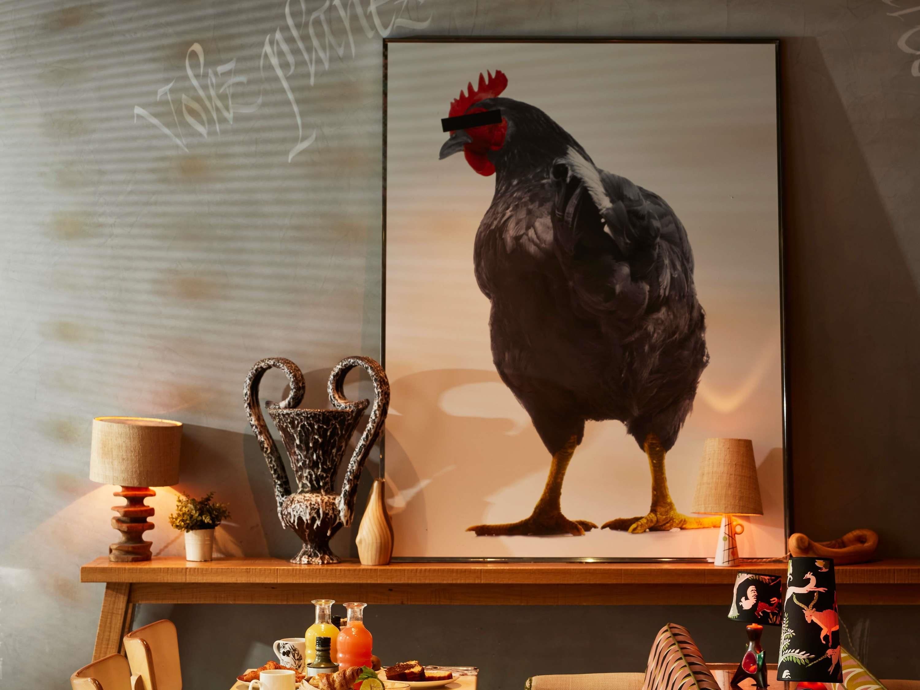 A framed rooster artwork on a wall, a wooden shelf with decorative items, and a table with breakfast items.