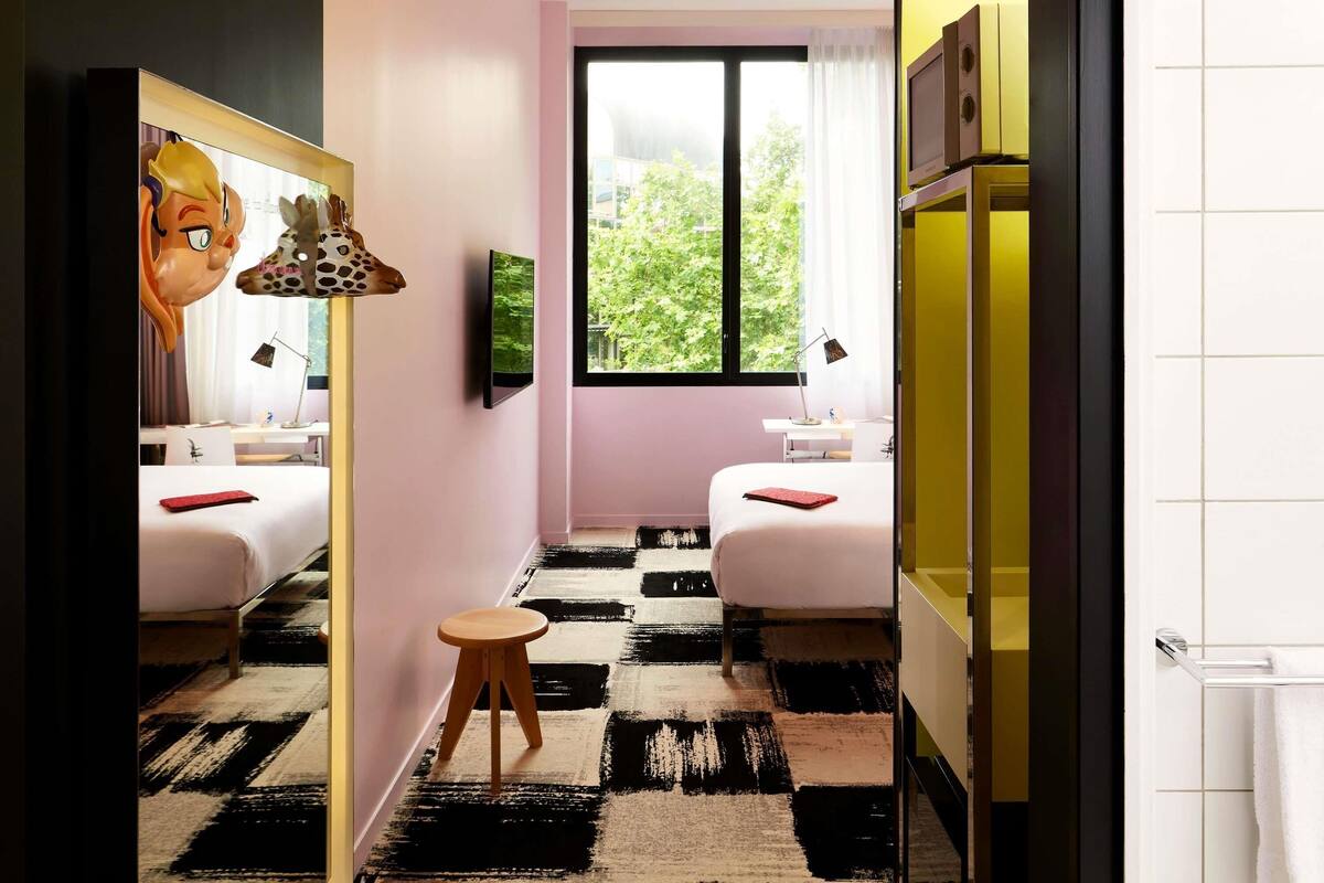 Een hotelkamer met een zwart-wit geruit vloerkleed, een bureau, een stoel, een bed, een televisie en een raam met uitzicht op bomen.
