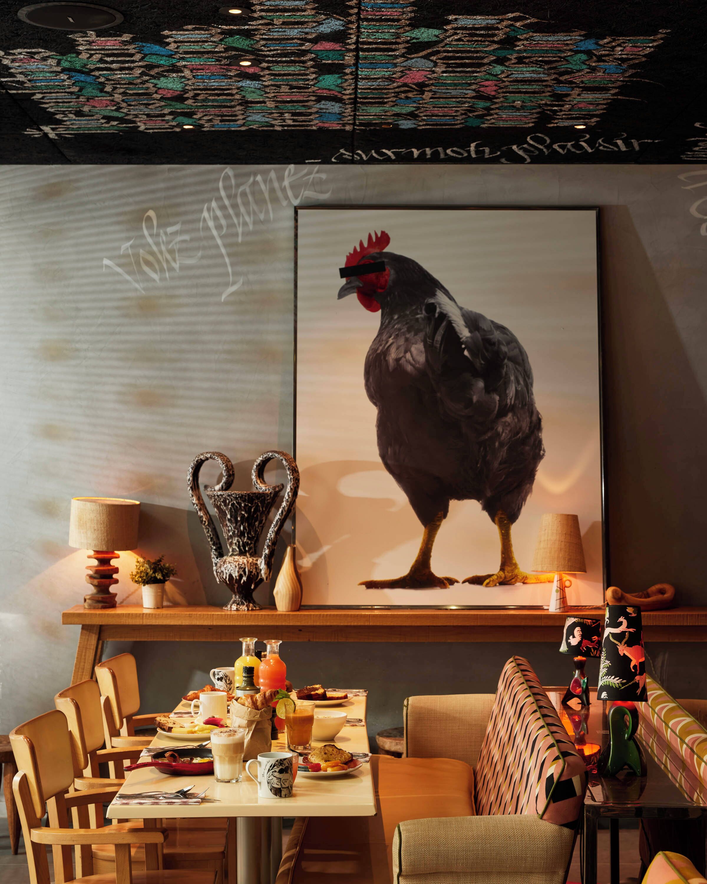A dining area with a table set for breakfast, a large framed rooster artwork on the wall, and a unique ceiling design.