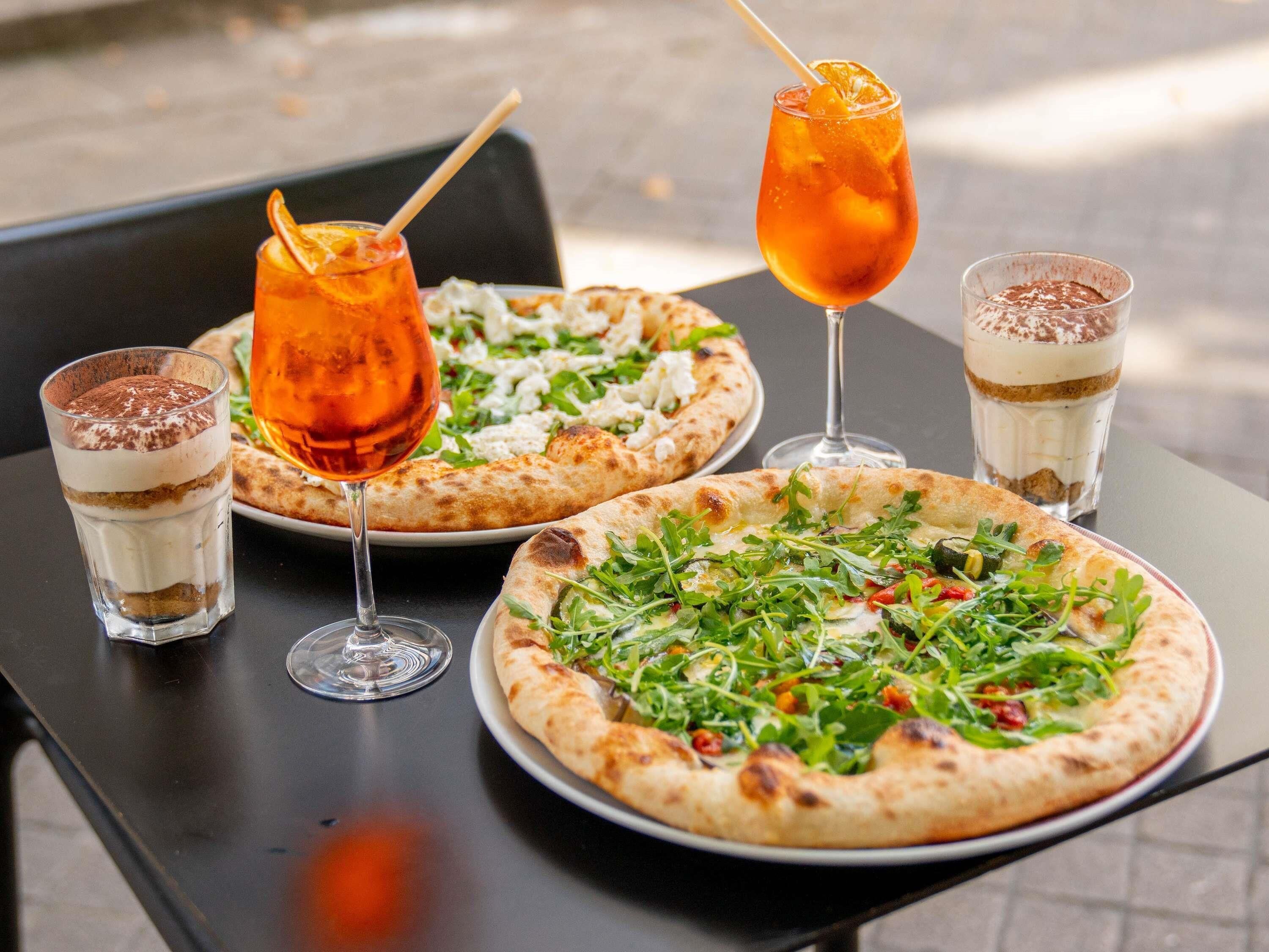 Una mesa con dos pizzas, un vaso de té helado y una bebida de postre.