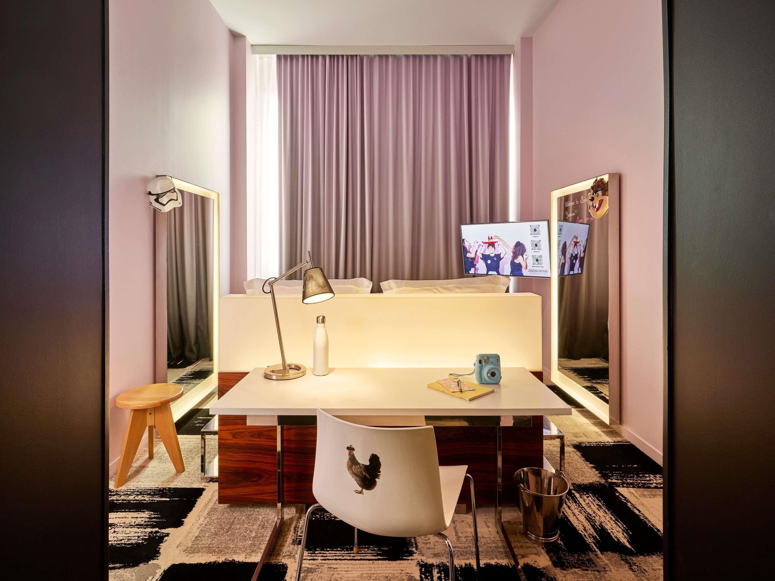 A hotel room with a desk, chair, lamp, bedside table, and a large mirror.