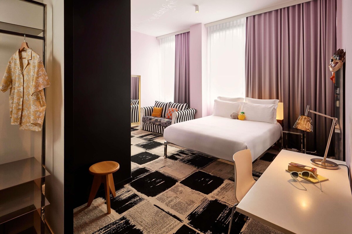 Een hotelkamer met een bed, een stoel, een klein tafeltje, een bureau, een spiegel en een kledingkast.