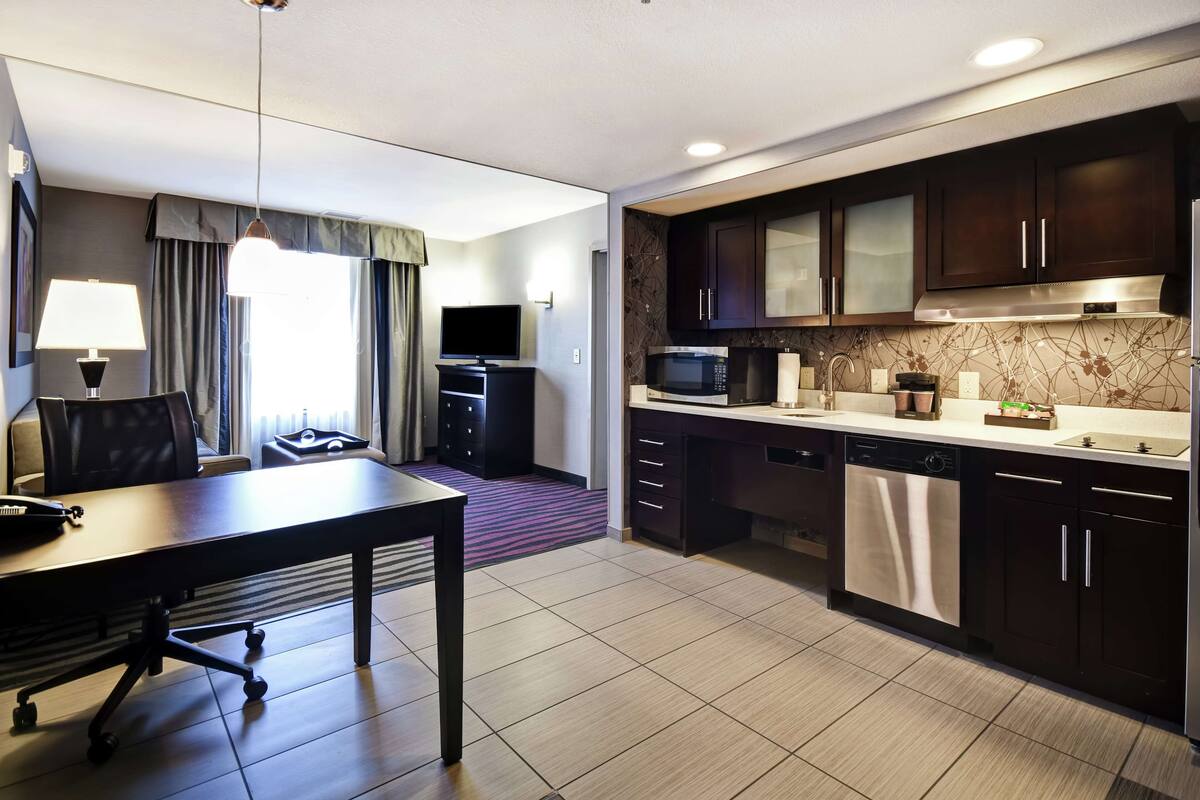 A modern hotel room with a dining area, a kitchen with dark cabinets and stainless steel appliances, and a large window with curtains.