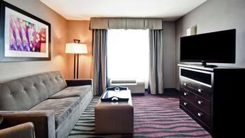 Une chambre d’hôtel avec un canapé gris, un téléviseur à écran plat, un petit pouf, une lampe et une grande fenêtre avec des rideaux.