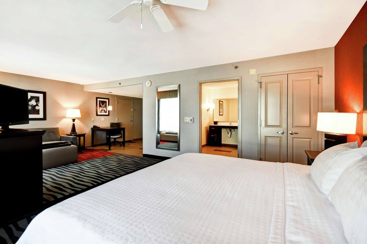 A hotel room with a large bed, a desk, a chair, a sofa, a mirror, and a wardrobe.