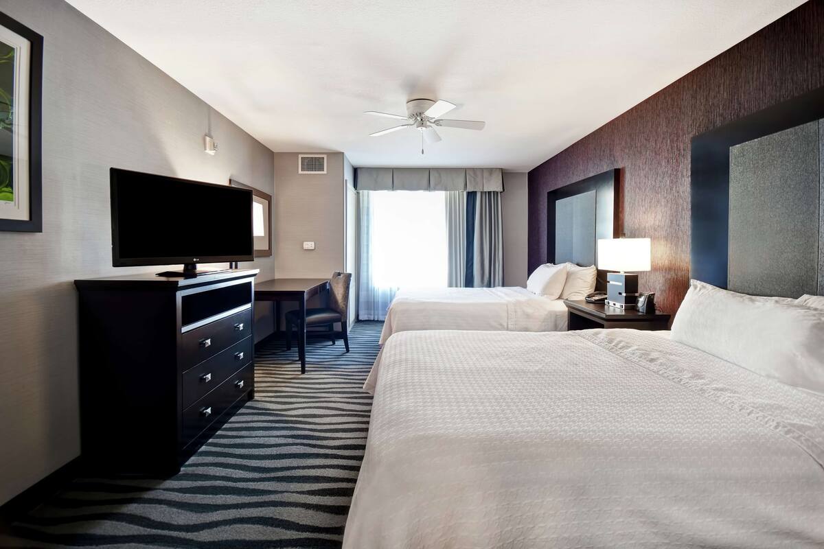 A hotel room with two beds, a flat-screen TV, a desk, and a ceiling fan.