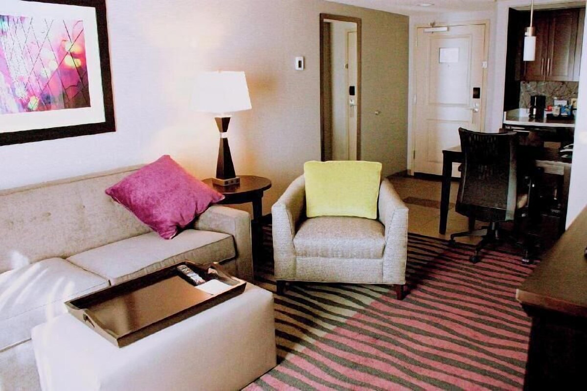 A hotel room with a sofa, armchair, a small table with a tray, a lamp, a kitchen area with cabinets, and a striped carpet.
