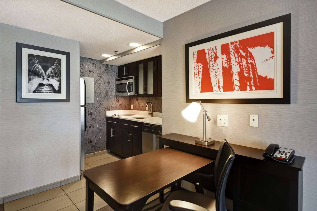 A modern kitchen with dark cabinets, a microwave, and a sink. There is a dining table with a chair and a desk with a lamp and a telephone.