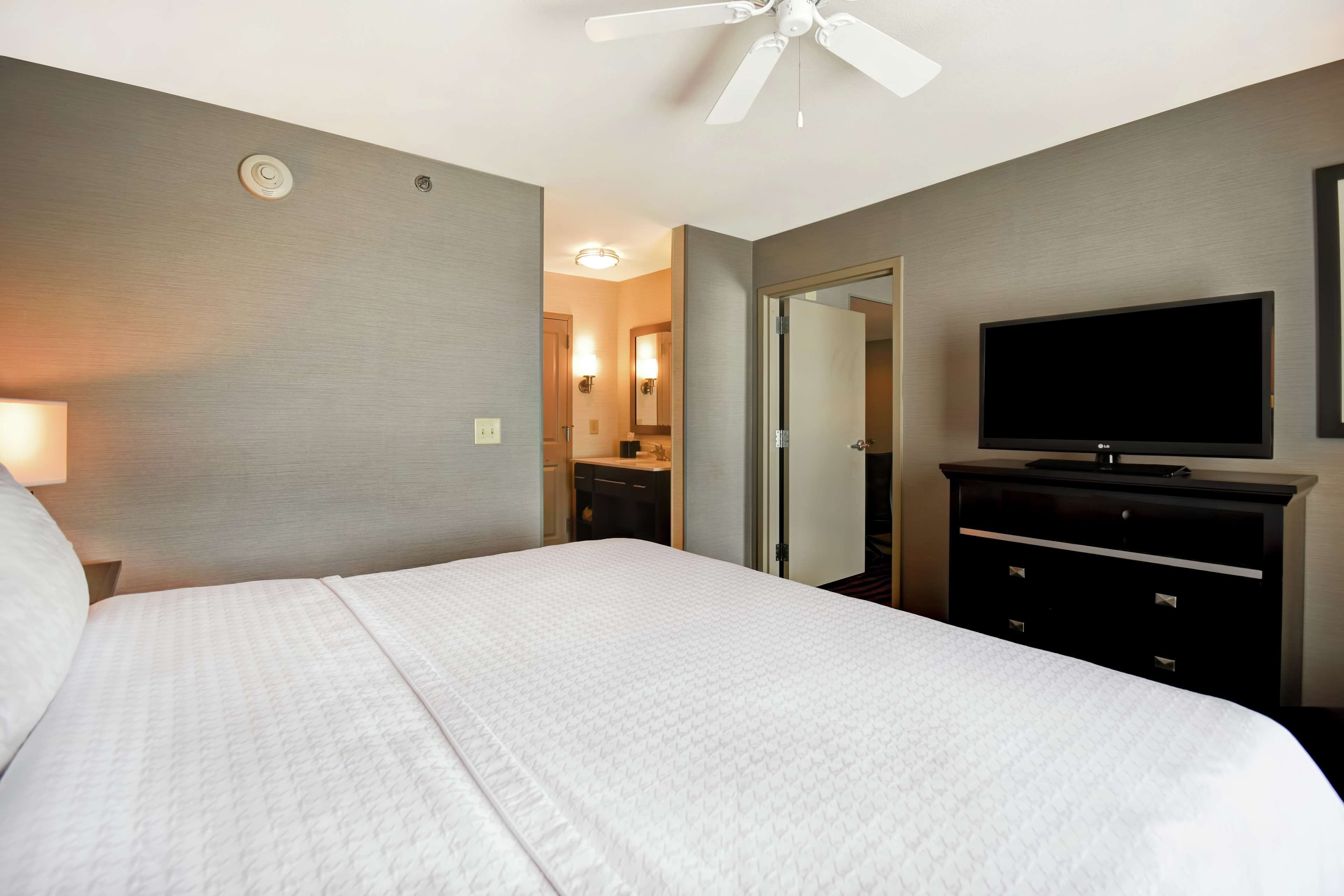 Une chambre d’hôtel dotée d’un grand lit, d’un téléviseur à écran plat, d’un ventilateur de plafond, d’une salle de bain avec un miroir et des luminaires, et d’une porte donnant sur une autre chambre.