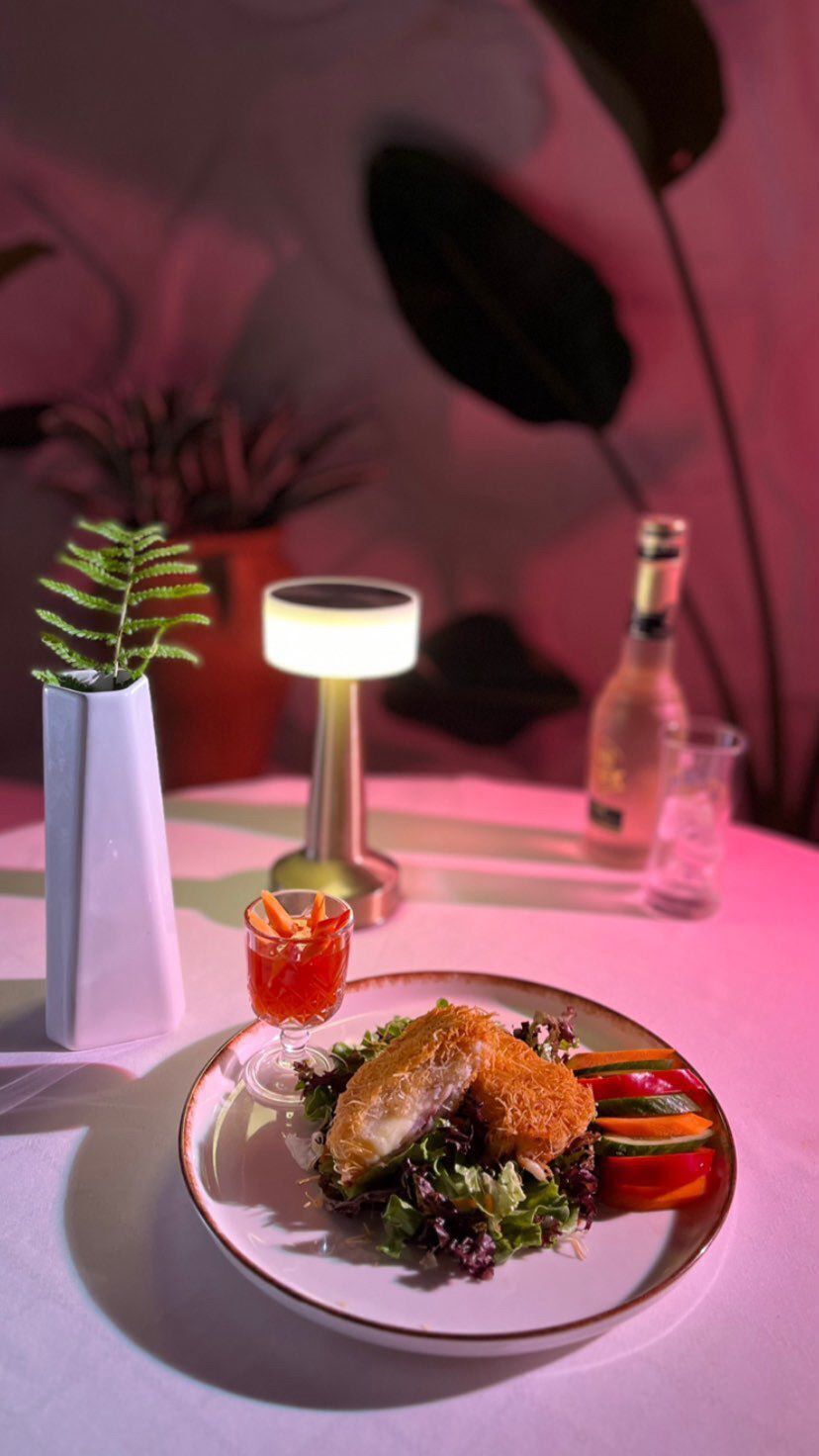 Une assiette de nourriture avec une salade, un petit verre d’une boisson rouge, une bouteille et une plante en pot, le tout posé sur une table.