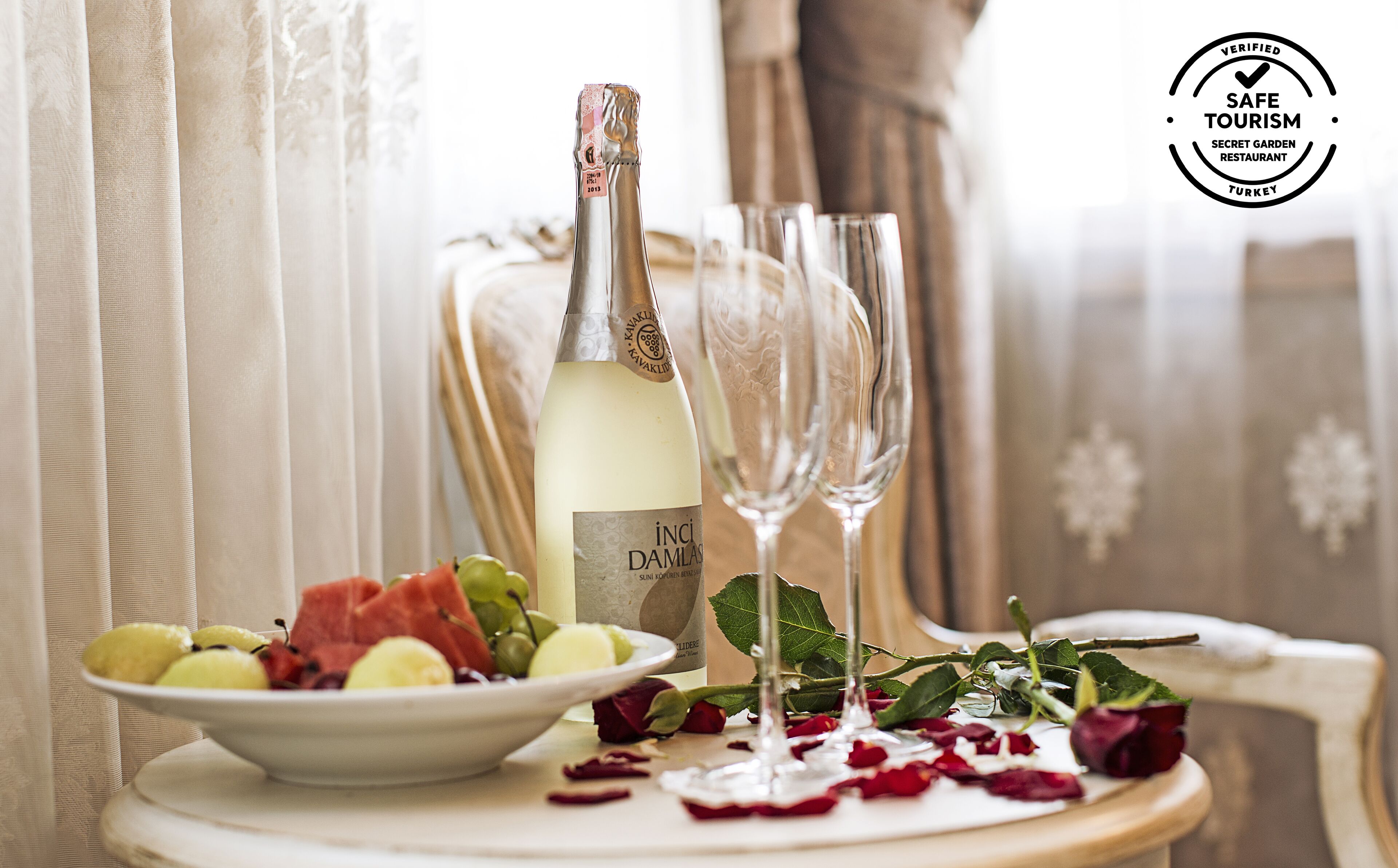 Una mesa con una botella de vino blanco, dos copas, un plato de fruta y pétalos de rosa.