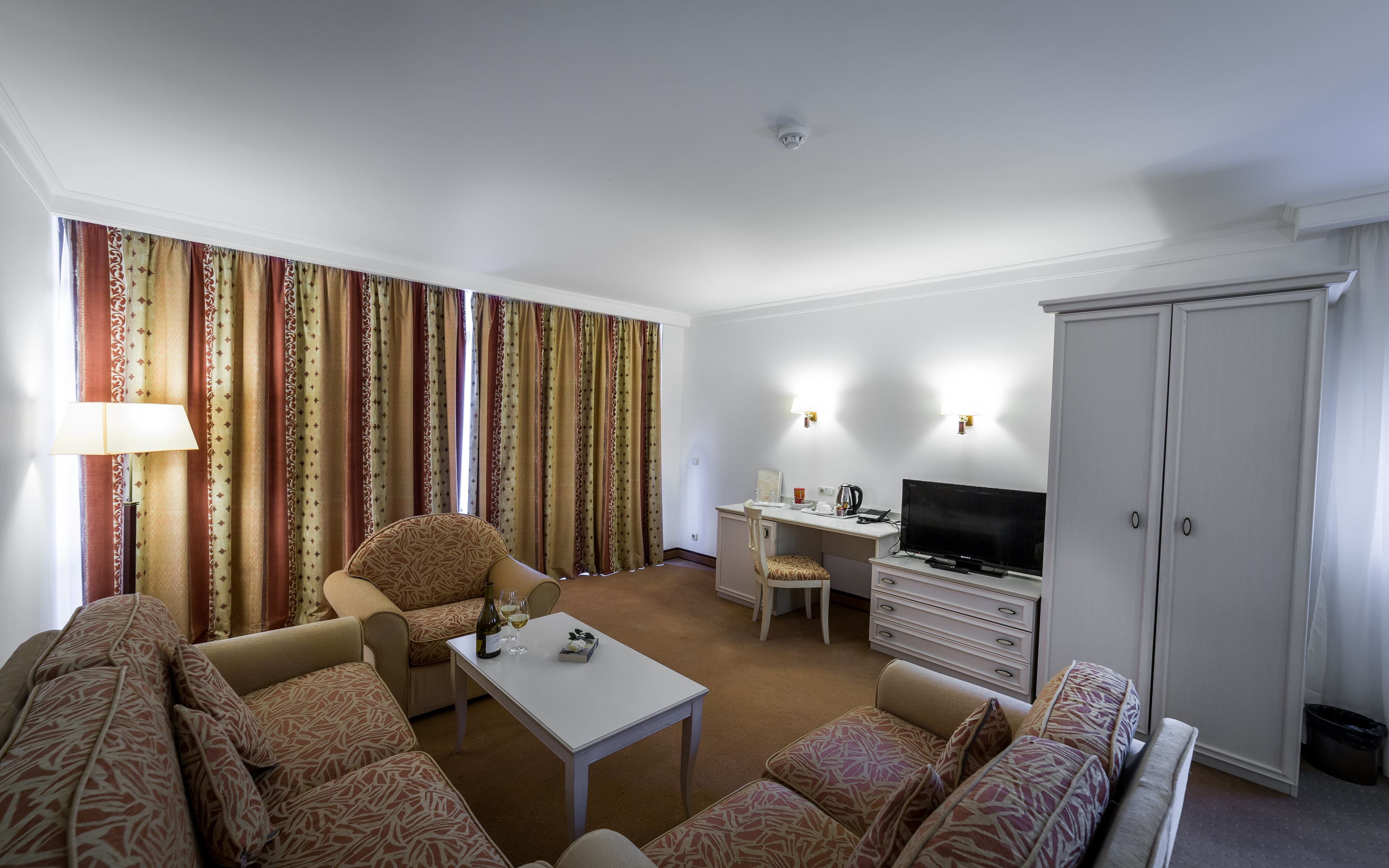 A hotel room with a sofa, armchair, dining table, TV, and wardrobe.