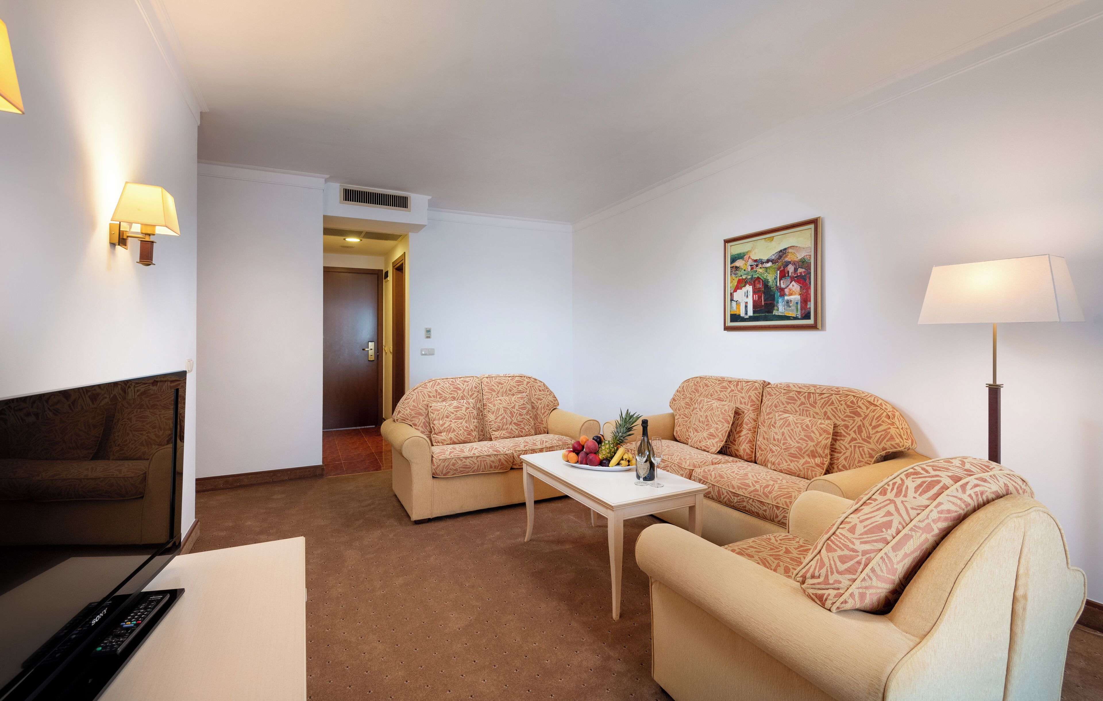 Une chambre d’hôtel avec un canapé, une table basse sur laquelle sont disposés des fruits, une lampe et un tableau au mur.