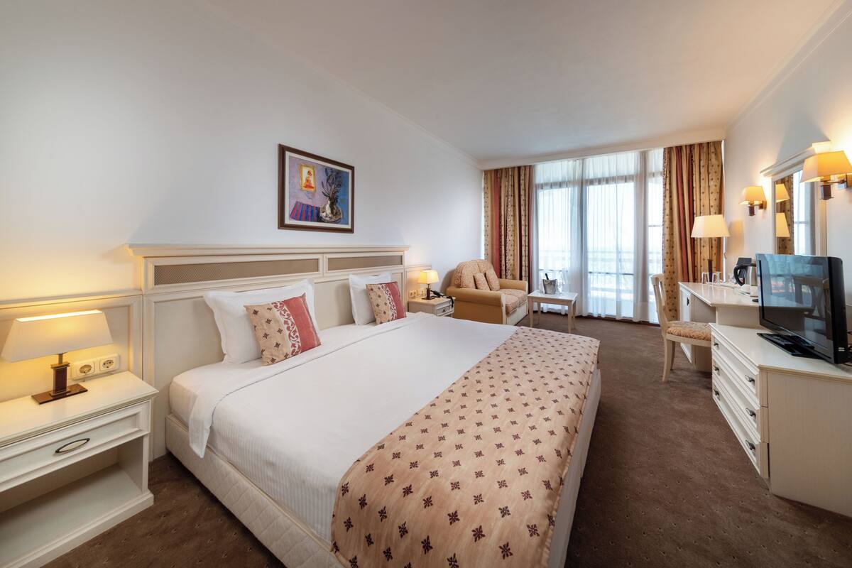 Una habitación de hotel con una cama grande, un escritorio, una silla, un televisor y un cuadro en la pared.