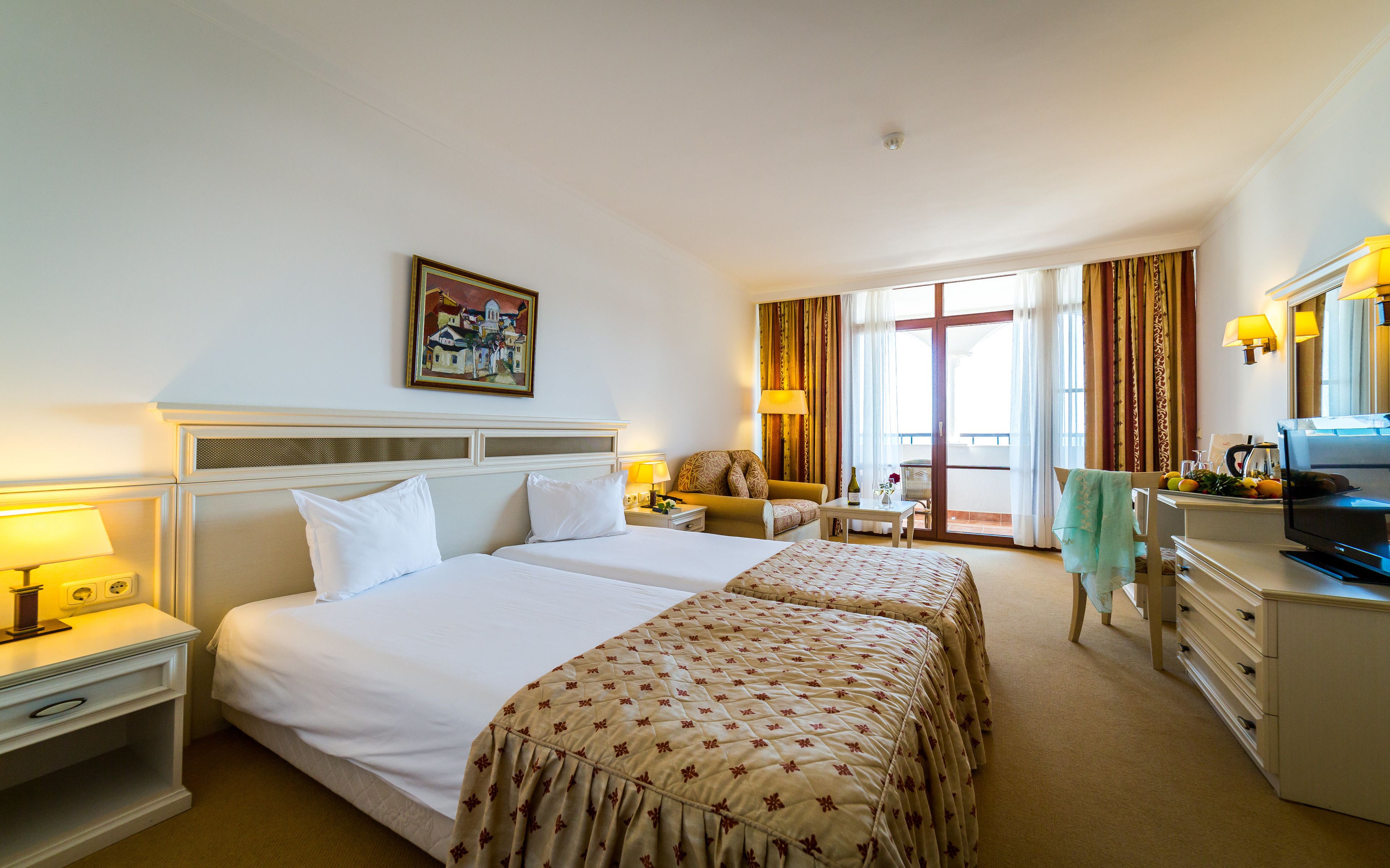 Ein Hotelzimmer mit zwei Betten, einem Schreibtisch, einem Sessel, einem Fernseher und einem Balkon mit Vorhängen.