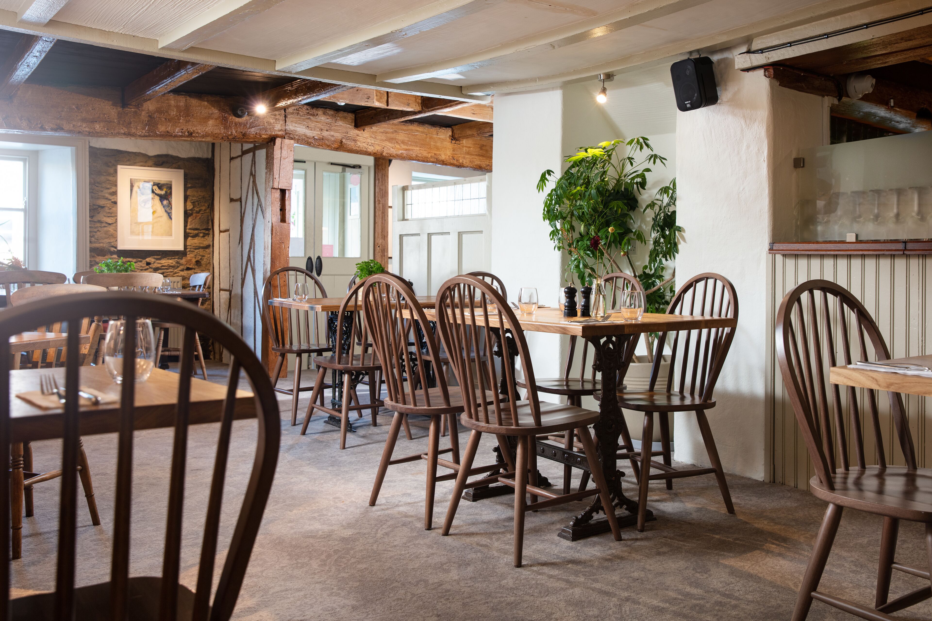 A rustic dining area with wooden tables and chairs, stone walls, and a large potted plant.