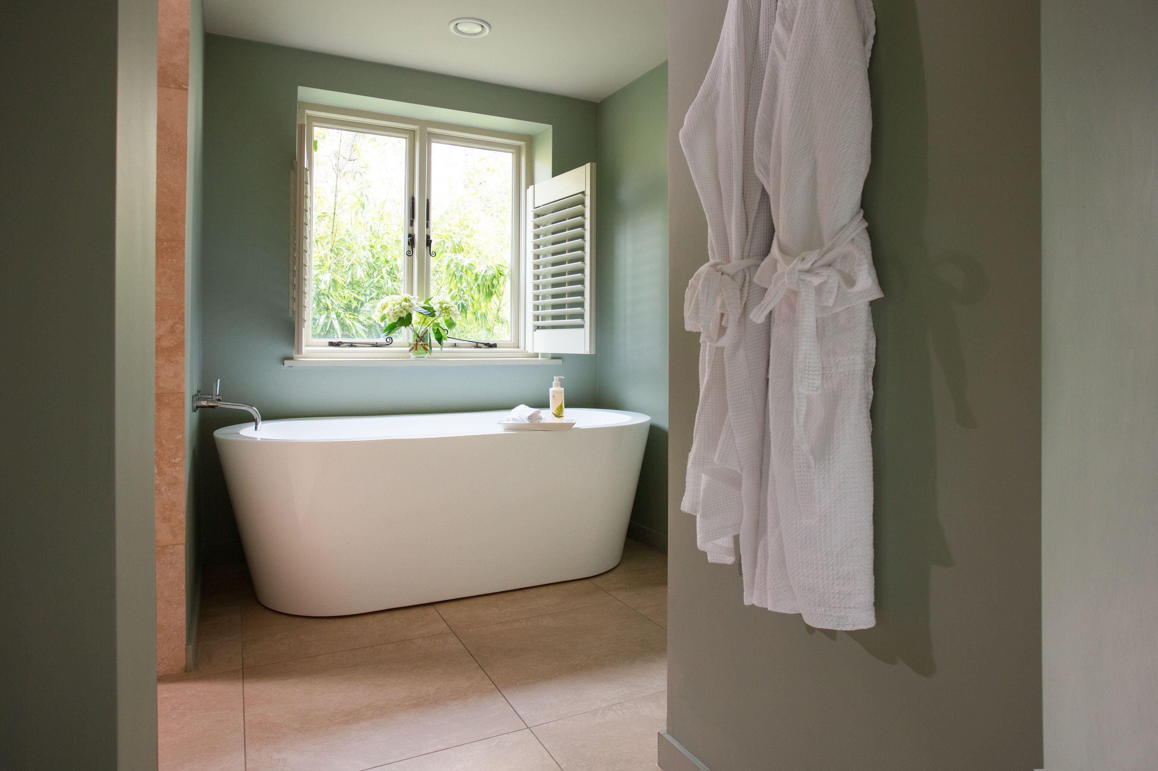 Une salle de bain avec une baignoire sur pieds, une fenêtre avec des volets et un peignoir blanc accroché au mur.