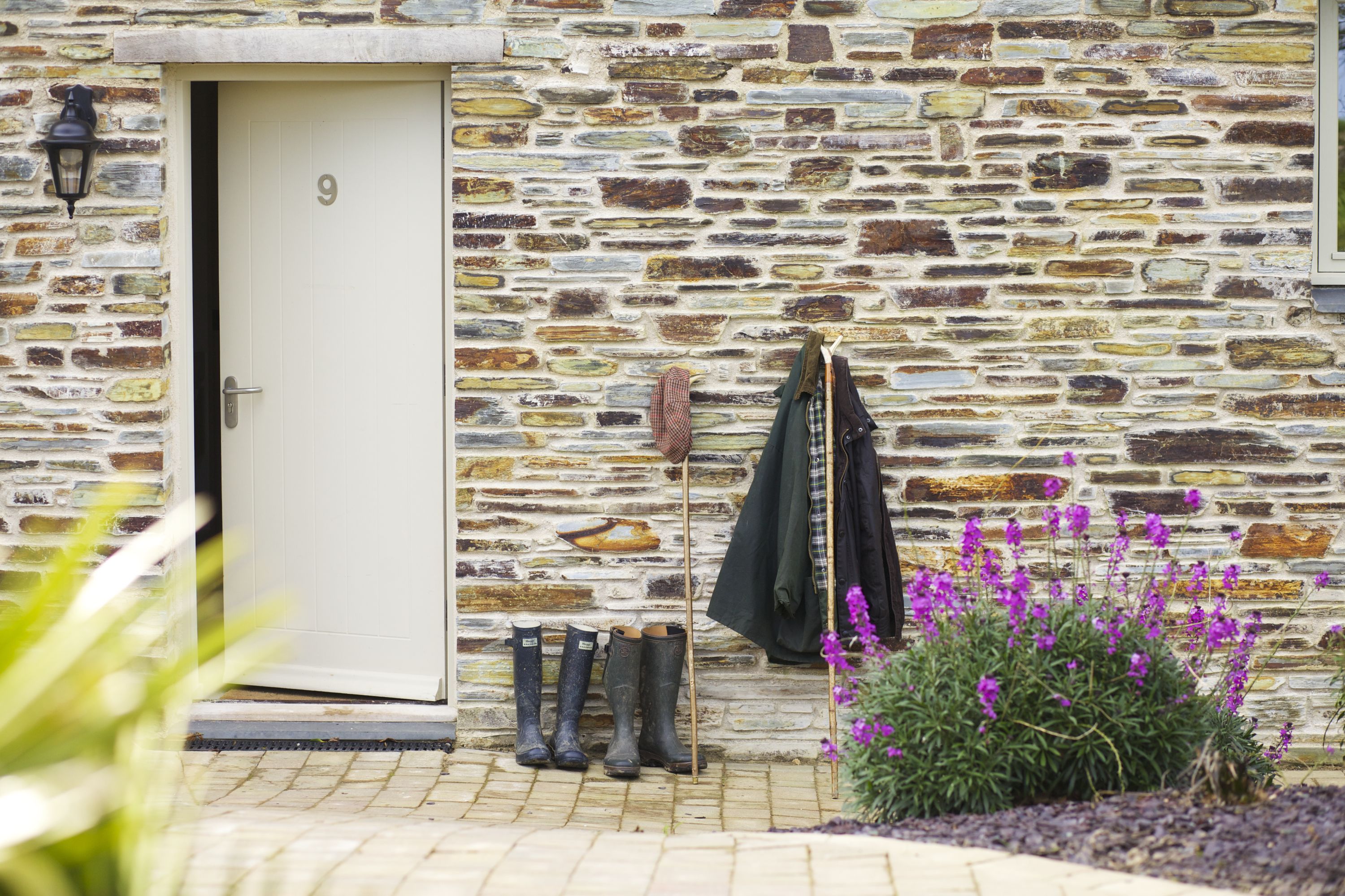 A stone building with a white door, boots, a coat, and purple flowers.