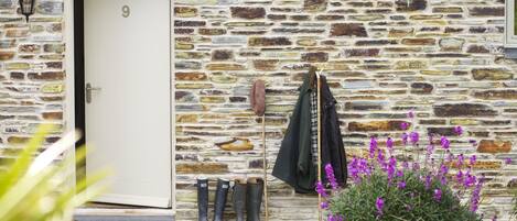 A stone building with a white door, boots, a coat, and purple flowers.