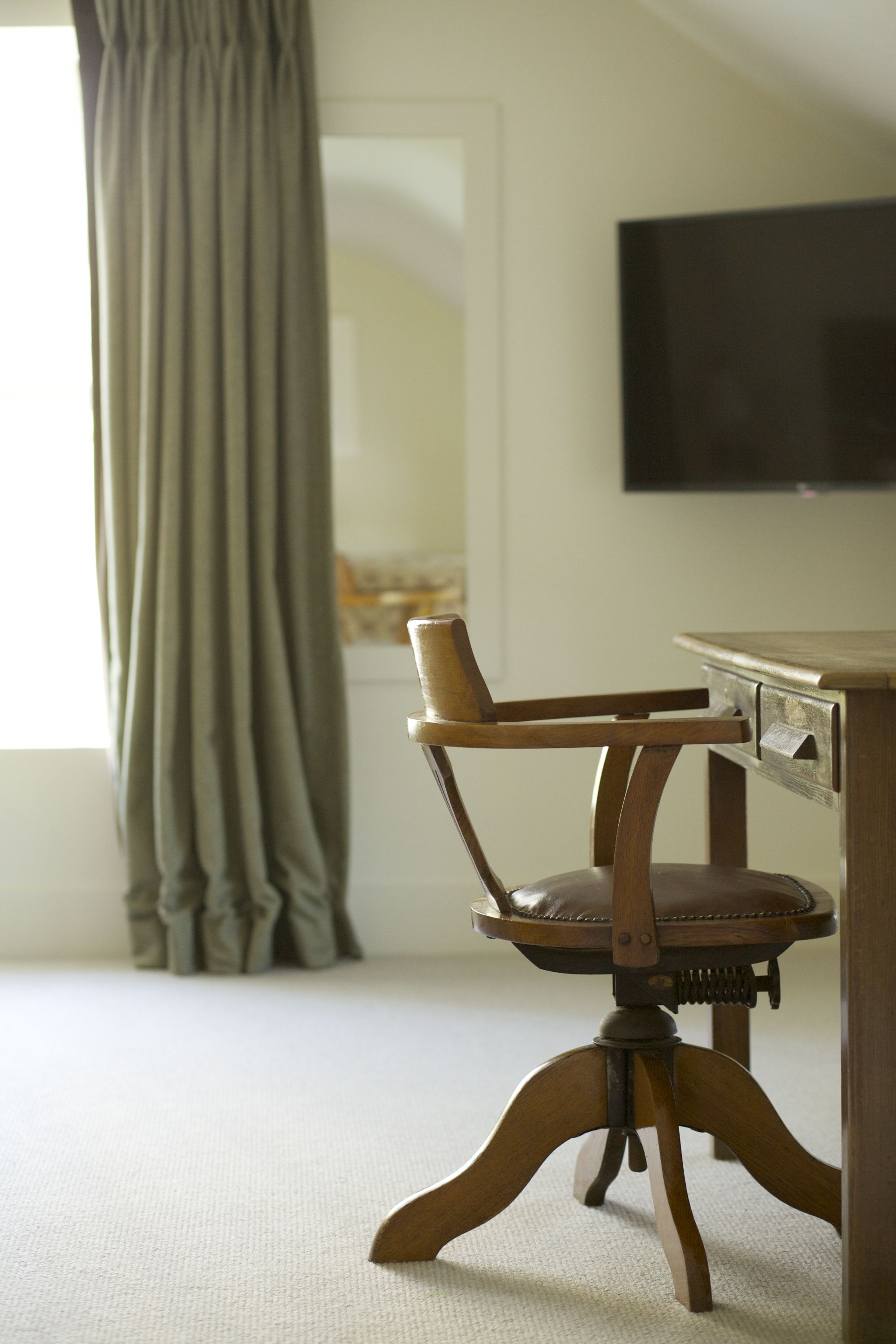 A wooden chair with a leather seat and a wooden table with drawers, positioned in a room with a light-colored carpet and a window with curtains.