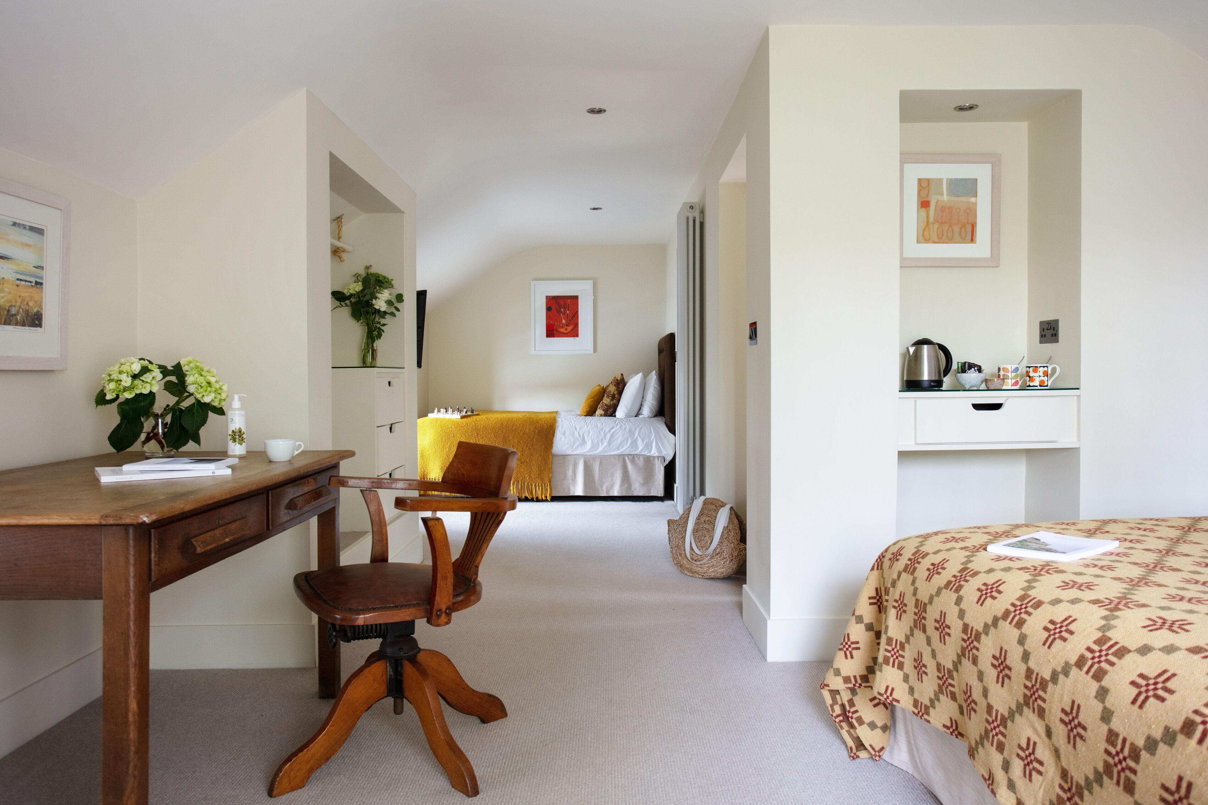 Habitación de hotel con un escritorio de madera, una silla, una cama con colcha estampada, una mesita de noche con hervidor y un cuadro en la pared.