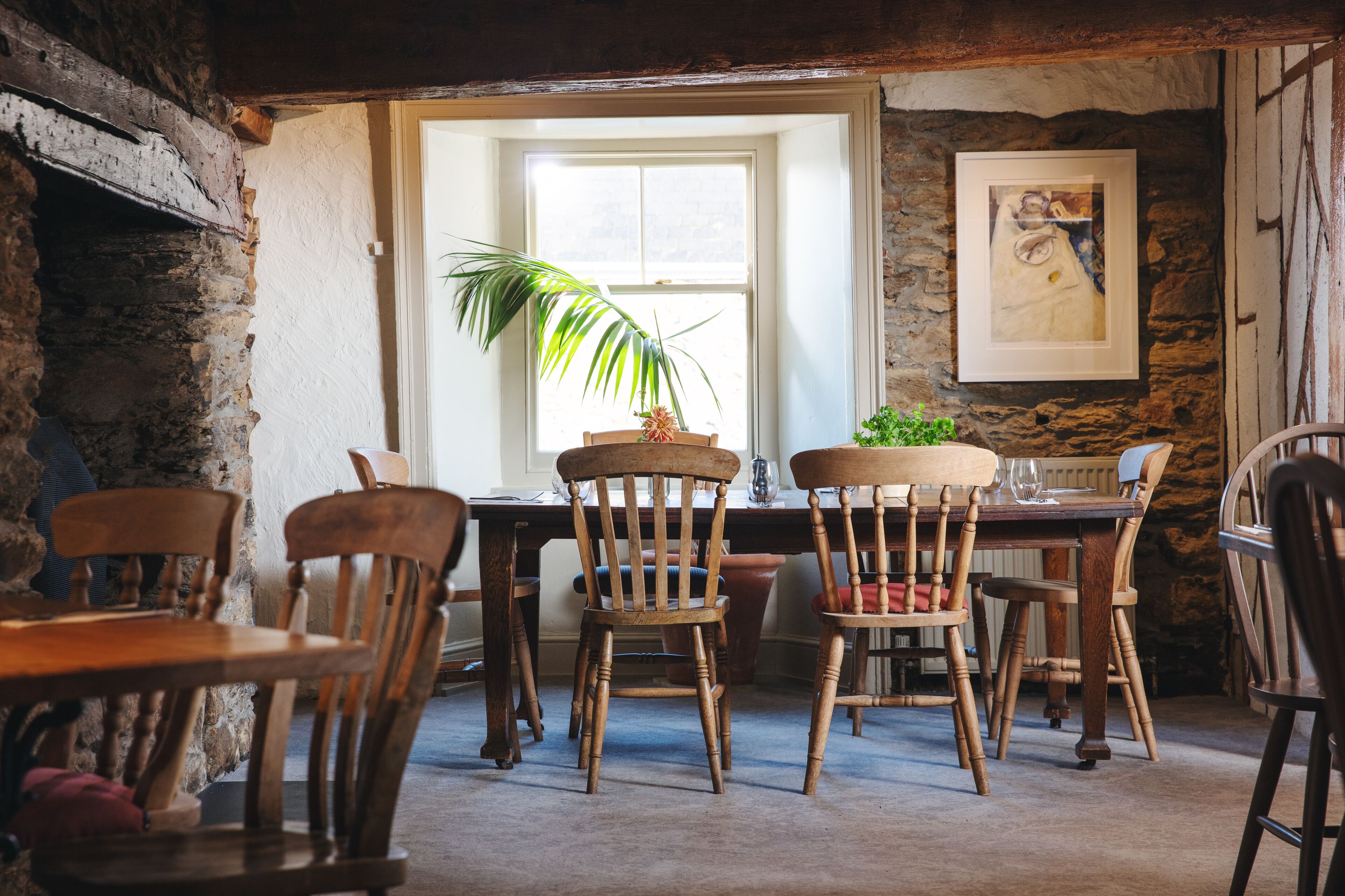 Een rustieke eethoek met houten stoelen en tafels, een stenen muur en een raam met een plant.