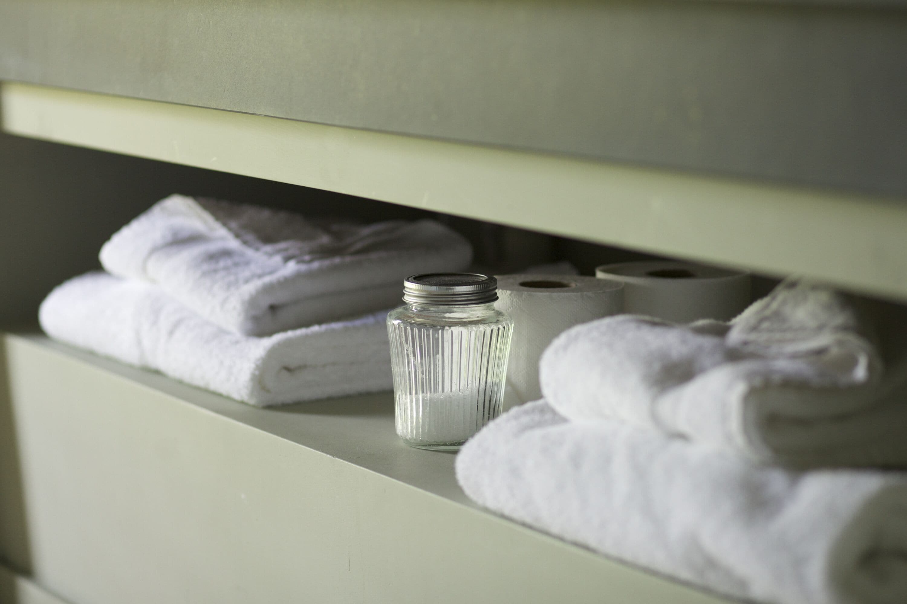 Une étagère avec des serviettes blanches soigneusement pliées et une bouteille en verre, qui contient probablement un produit de nettoyage.