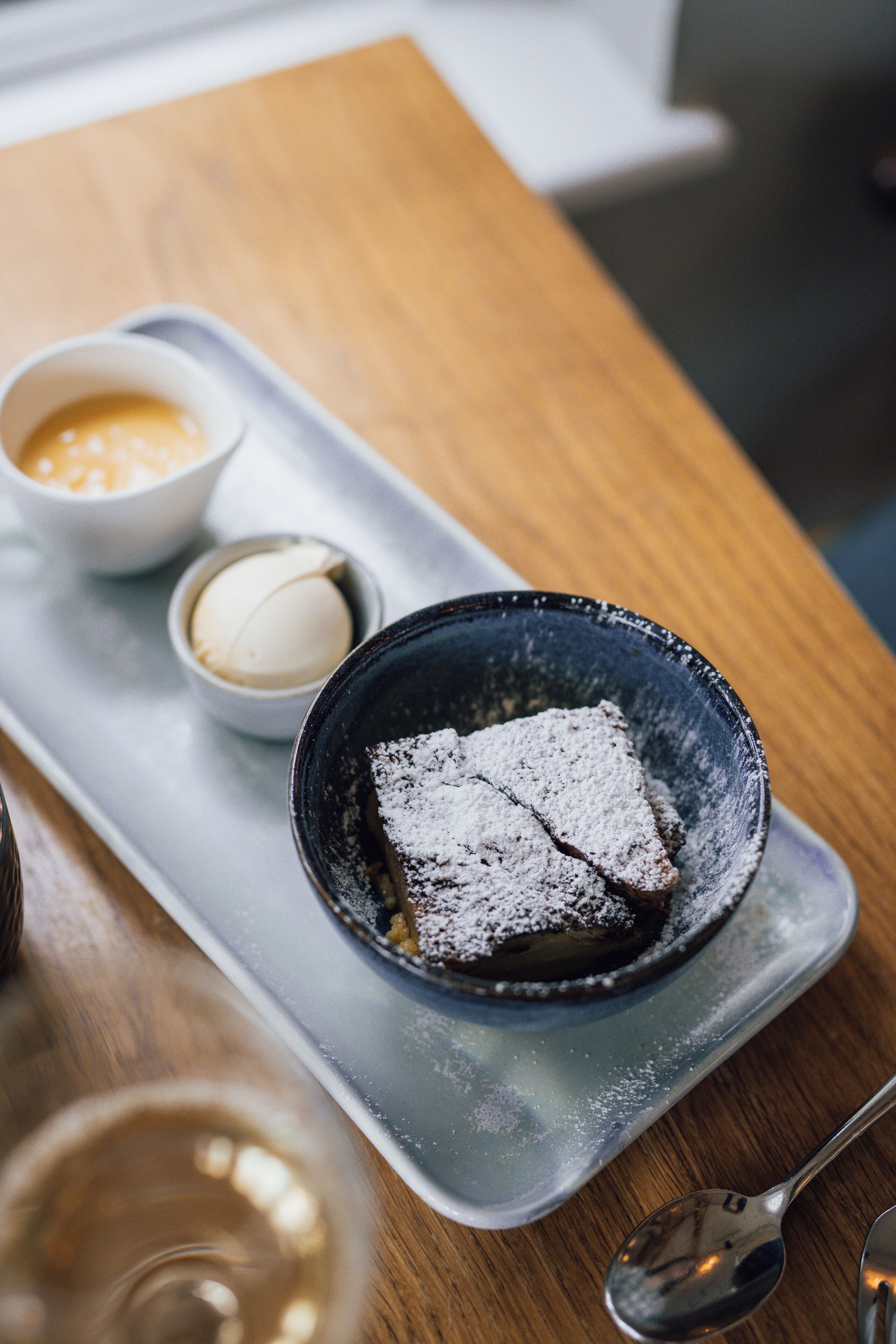 A dessert tray with a chocolate dessert dusted with powdered sugar, accompanied by a small bowl of whipped cream and a small bowl of orange sauce.