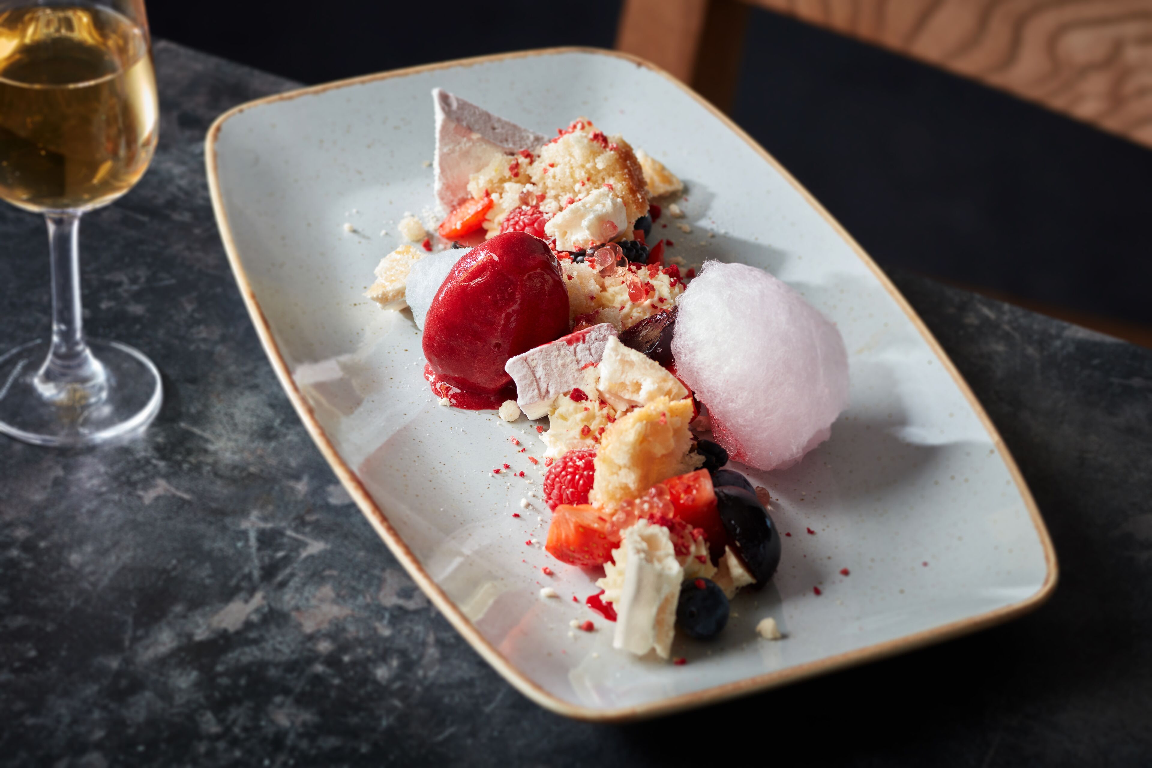 Un plato de postre con diversas frutas y una copa de vino blanco sobre una mesa de mármol oscuro.