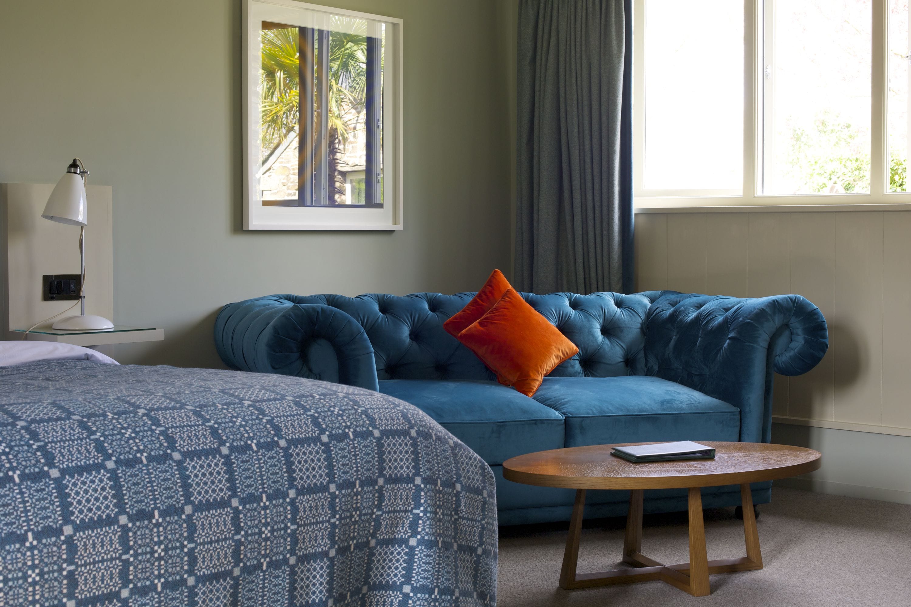 A room with a blue sofa, a round wooden table, a patterned bedspread, a framed picture on the wall, and a window with curtains.