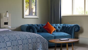 A room with a blue sofa, a round wooden table, a patterned bedspread, a framed picture on the wall, and a window with curtains.