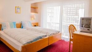 A hotel room with a double bed, a desk with a television, a chair, a window with sheer curtains, and a framed picture on the wall. - Hotel Heilquelle (Leukerbad)