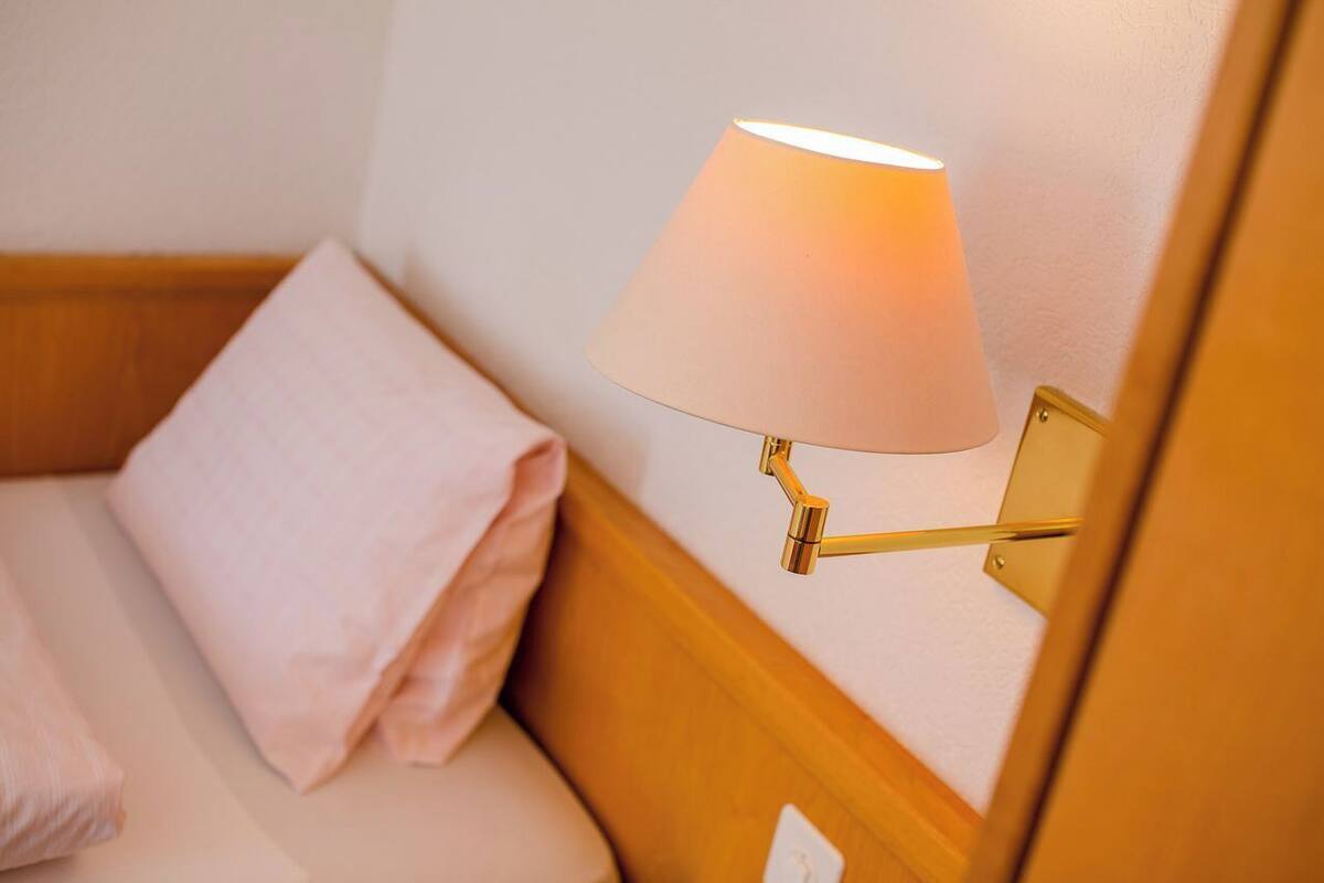 Un lit bien fait, avec un oreiller rose et une lampe fixée au mur, dotée d’un abat-jour blanc.