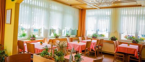 A restaurant with multiple tables, each covered with a pink tablecloth. There are potted plants on the tables and along the windows. The room has wooden floors and a chandelier.