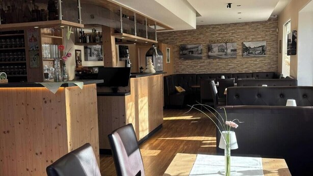 A modern bar area with wooden furniture, a seating area with a black couch, and a wall with framed pictures. - Kräuterhotel Heidejäger (Rotenburg)